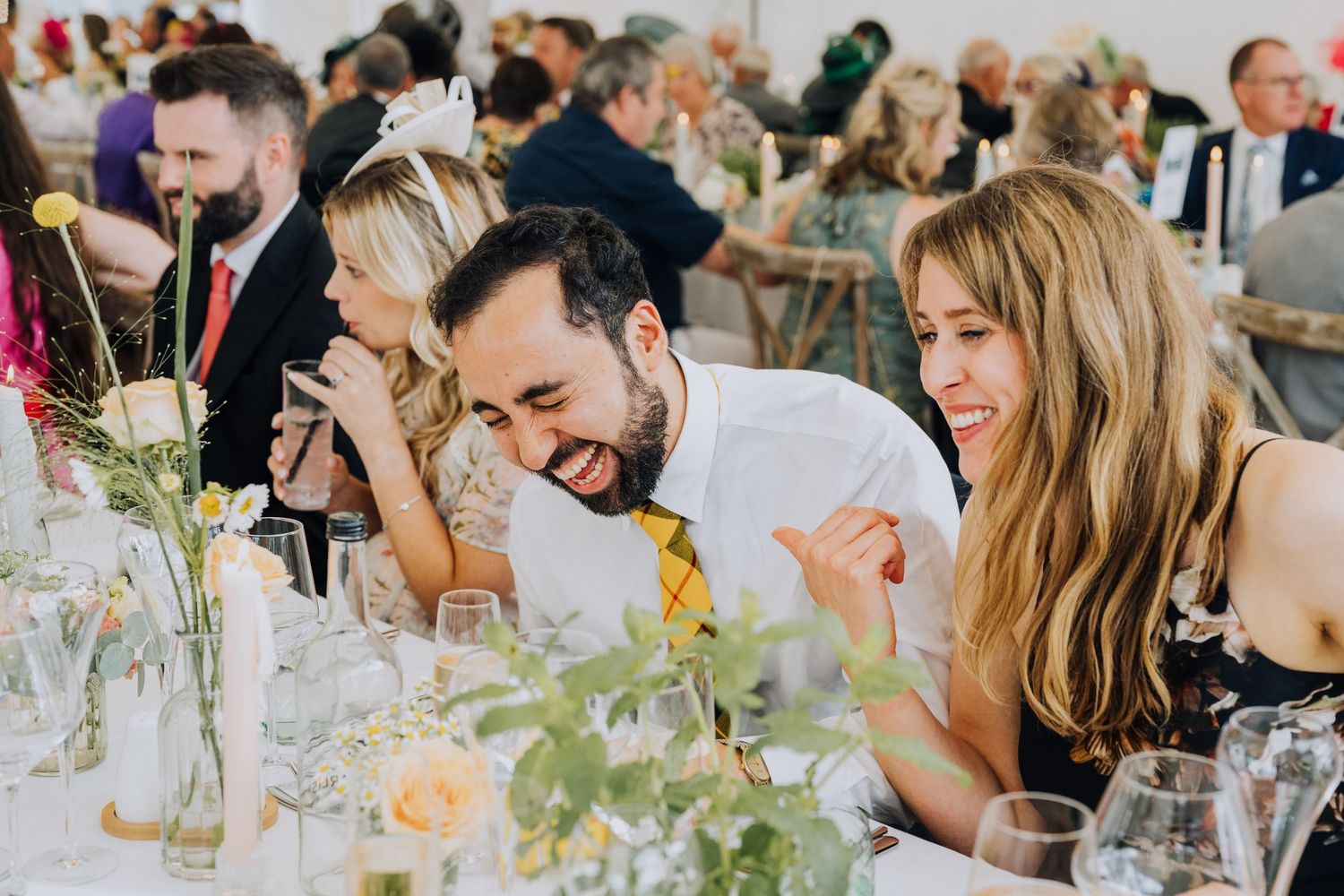 Wedding guests laughing and enjoying conversation at a bright, flower-decorated reception dinner table.