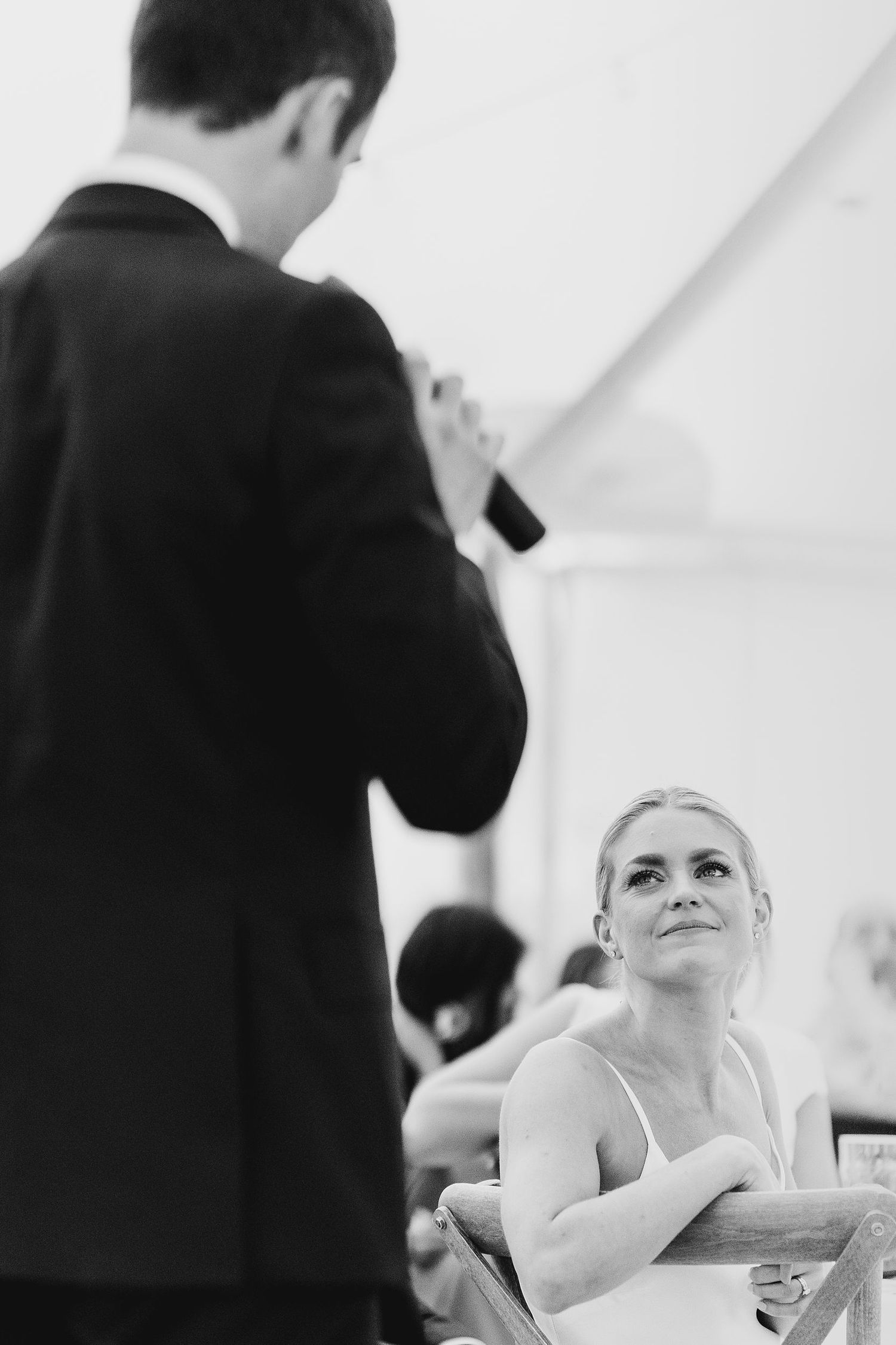 Bride gazing lovingly at groom delivering wedding speech at reception in black and white photo.
