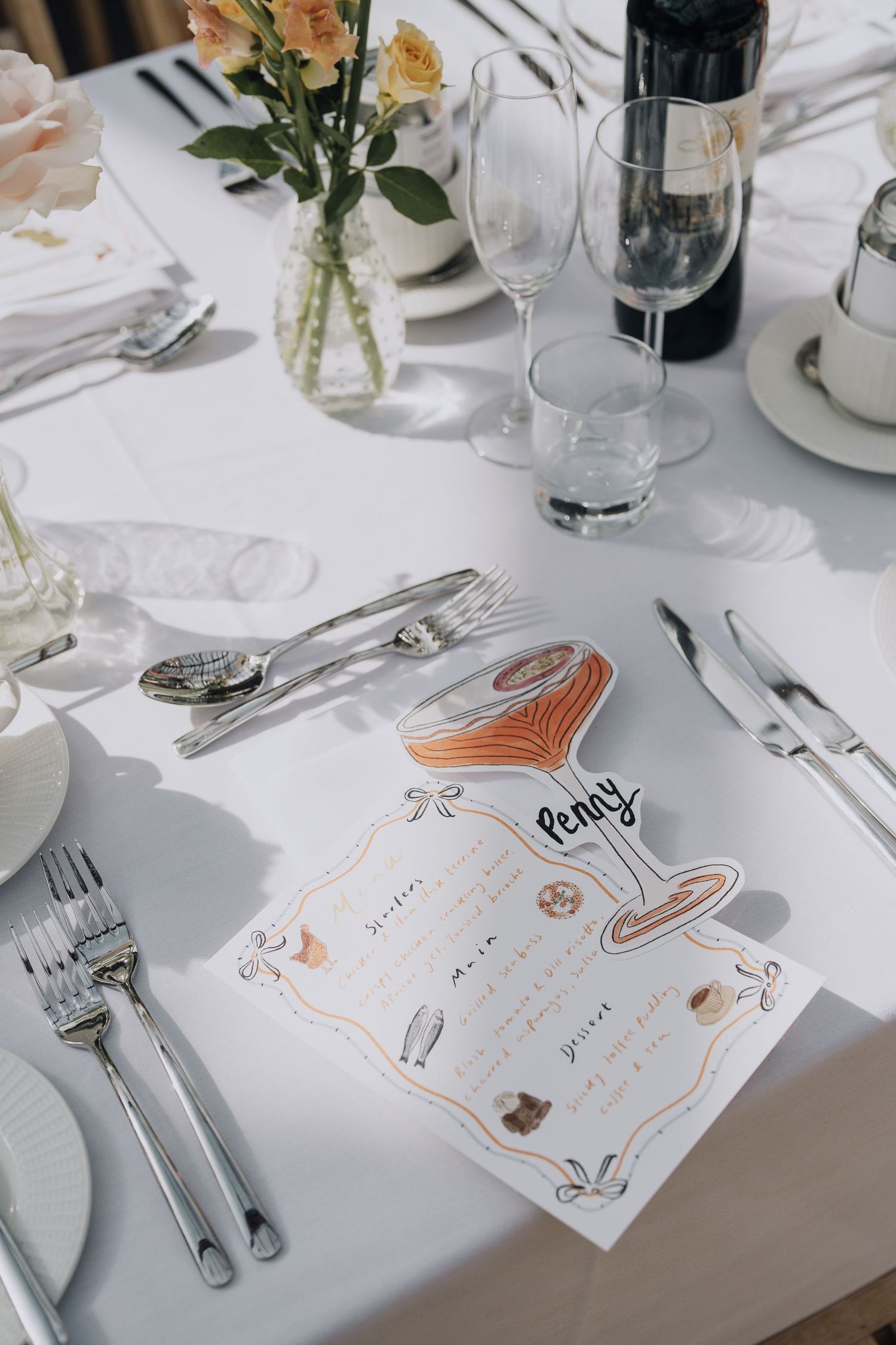 Elegant wedding place setting with illustrated cocktail menu card named Penny on a white tablecloth.