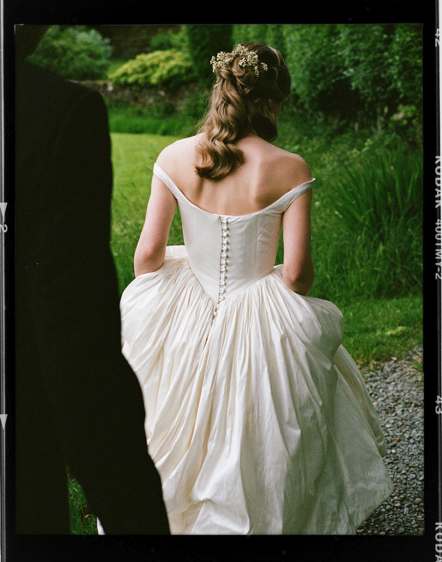 Bride in off-shoulder white wedding gown with corset back and floral hair accessory walks through lush garden.