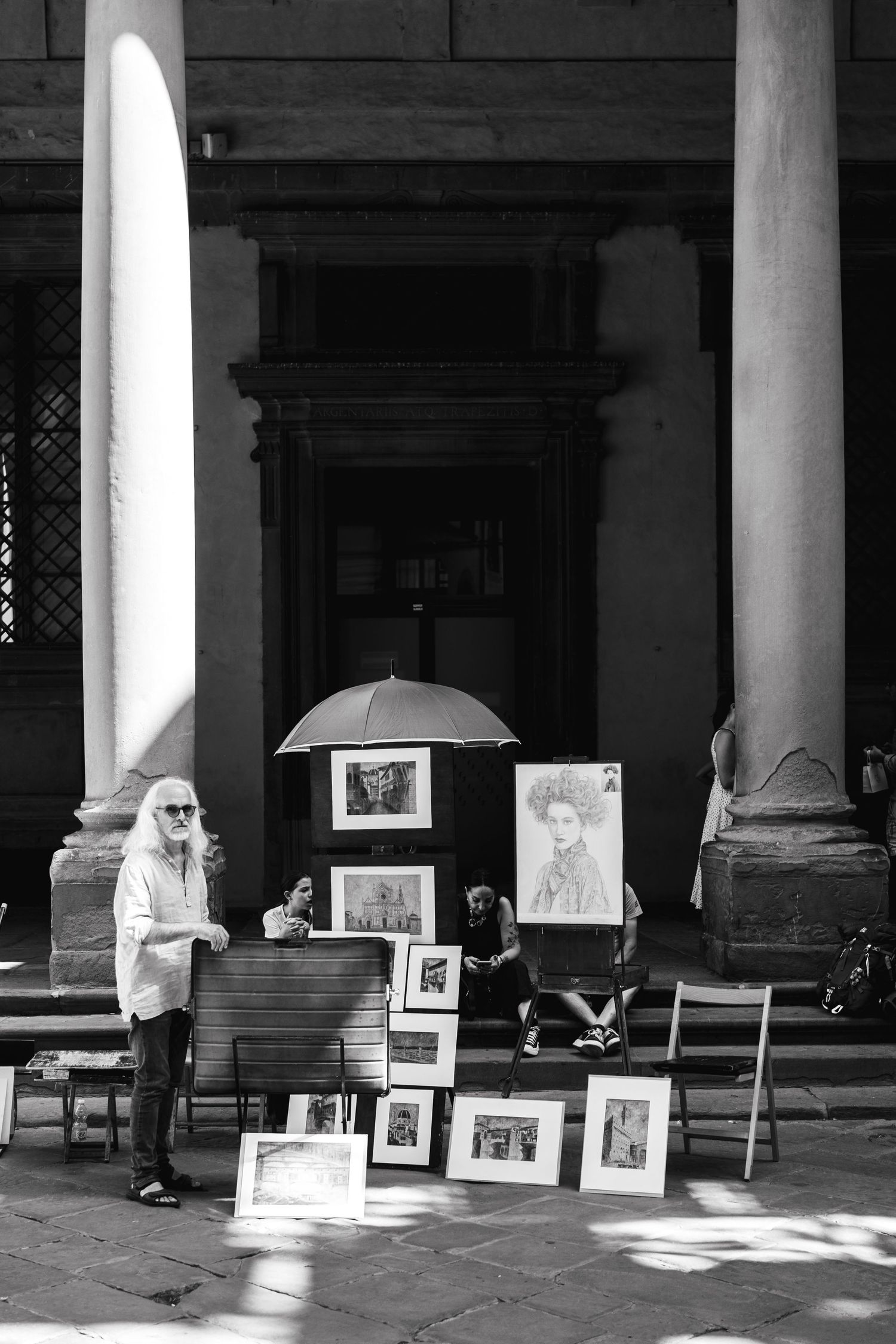 A street artist displaying framed artwork under a classical archway in black and white.