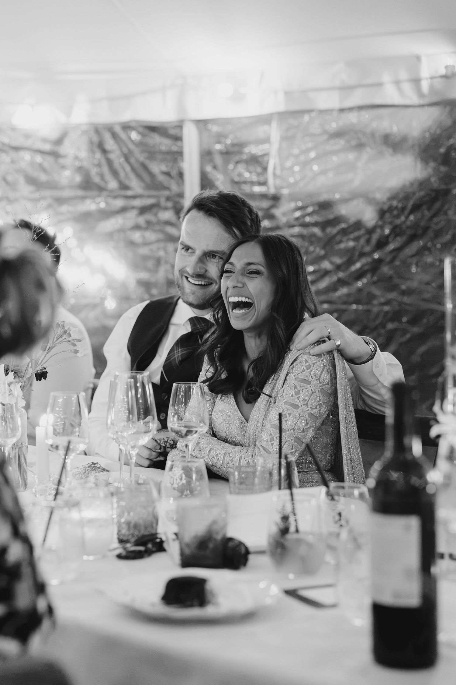 Laughing bride and groom embrace at wedding reception dinner table in black and white photo.
