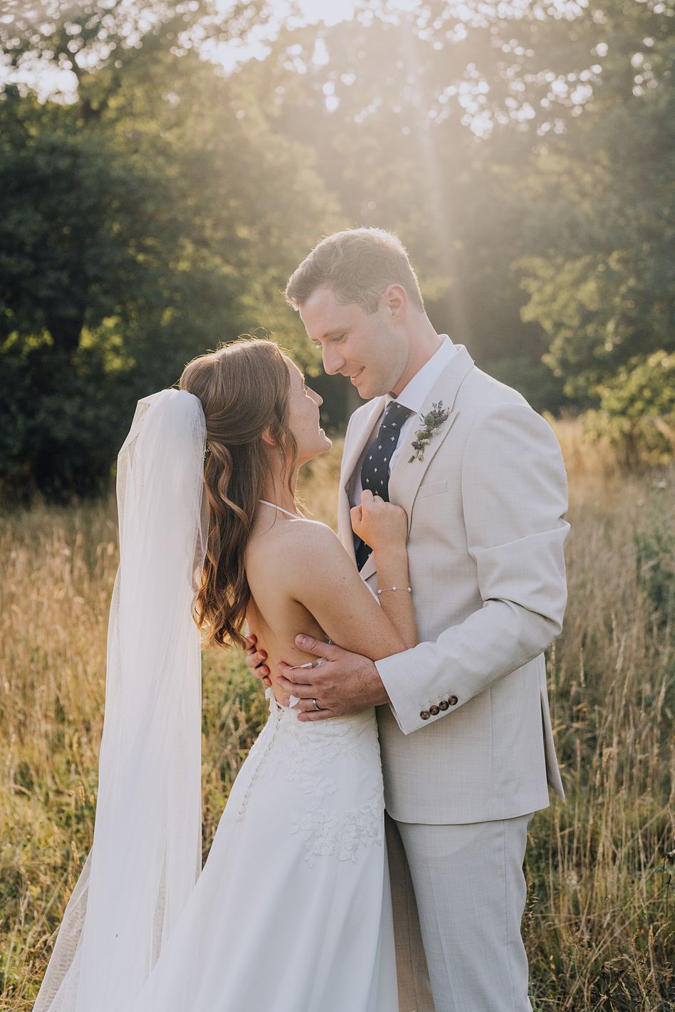 Bride and groom embrace in golden hour sunlight in a meadow, sharing a romantic moment.