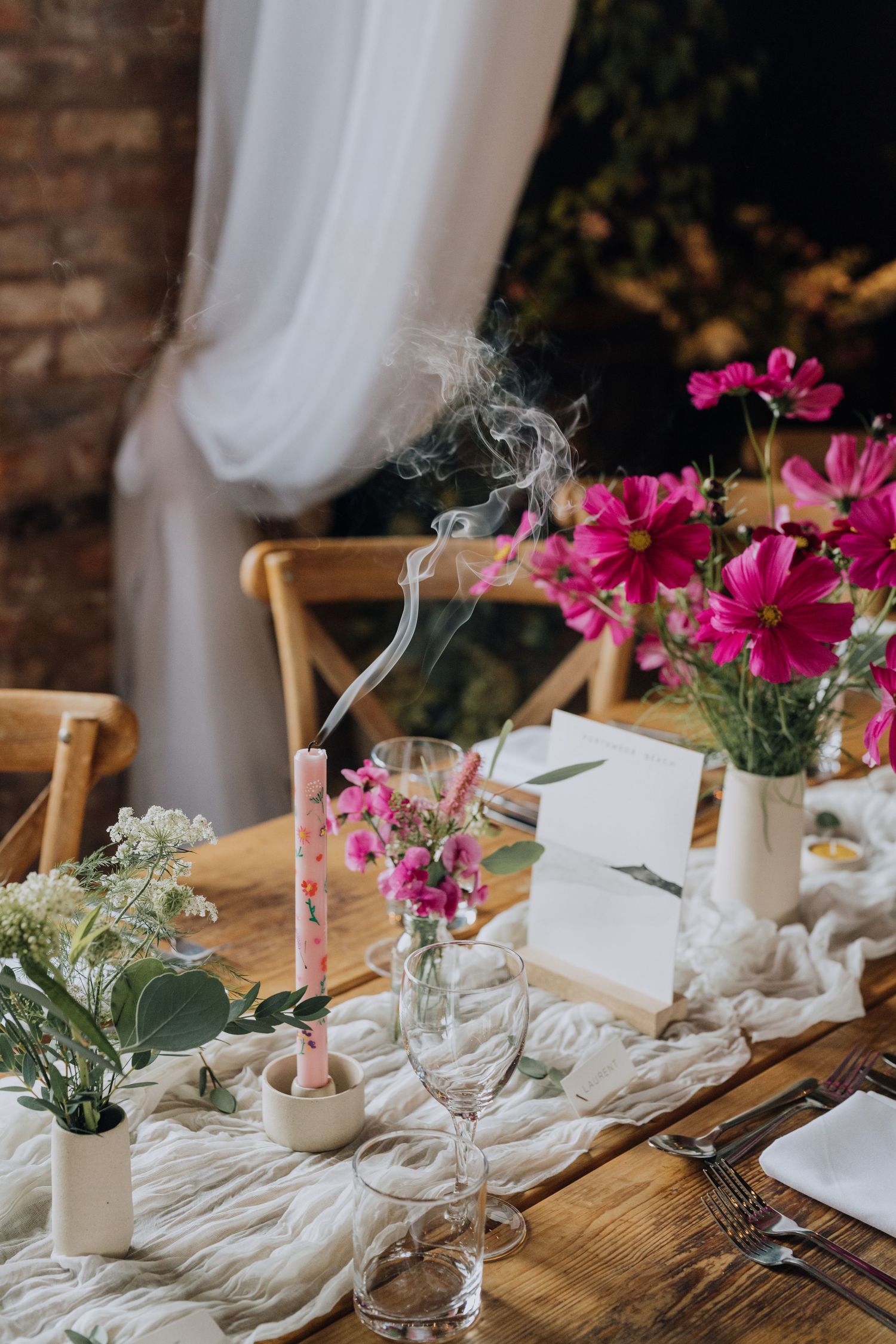 Elegant wedding table setting with pink cosmos flowers, a smoking candle, wine glasses, and white linen runner.