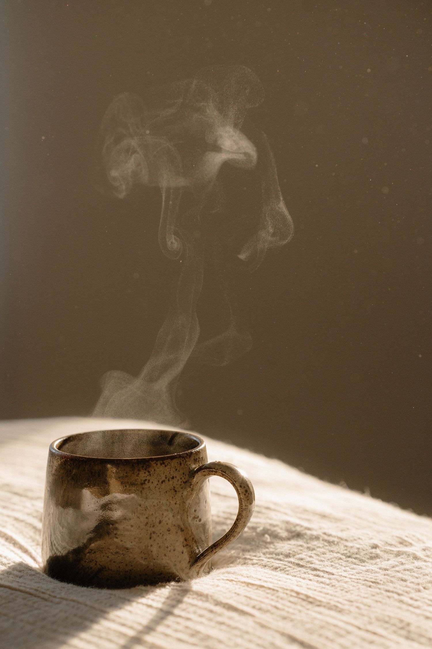 A steaming rustic ceramic mug on a textured white surface with warm sunlight casting soft shadows.