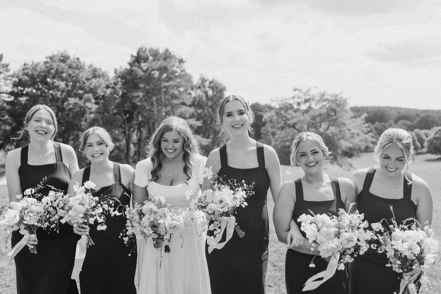 Bride and bridesmaids in black dresses holding wildflower bouquets laughing together outdoors in black and white photo.