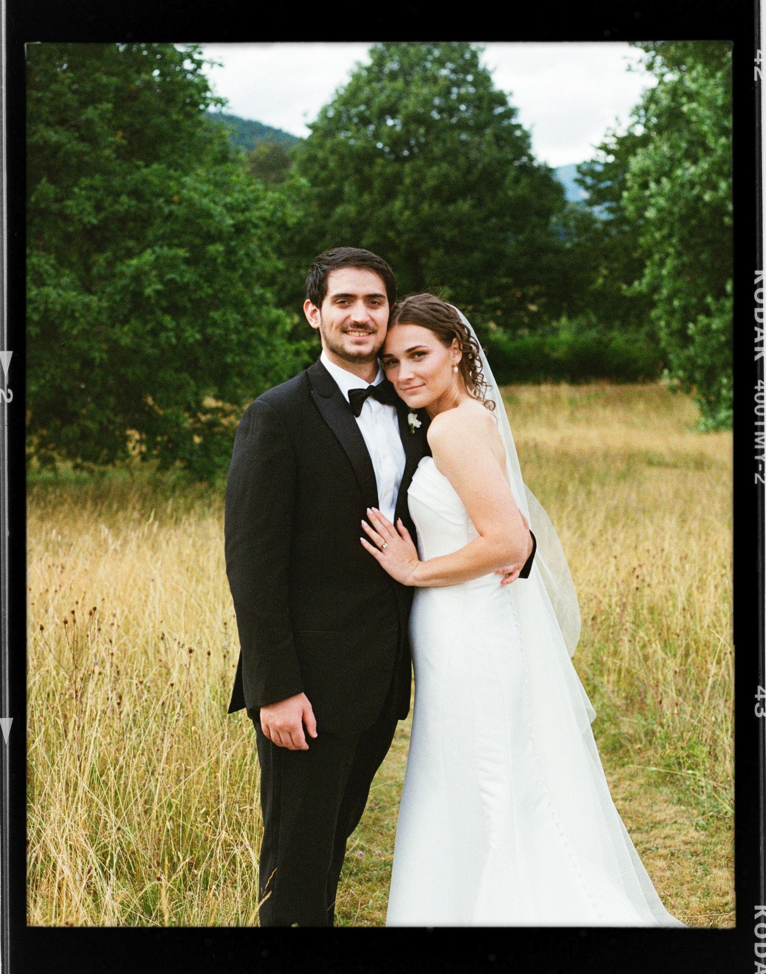 Bride and groom posing in a golden field with lush green trees and mountains in the background on their wedding day.
