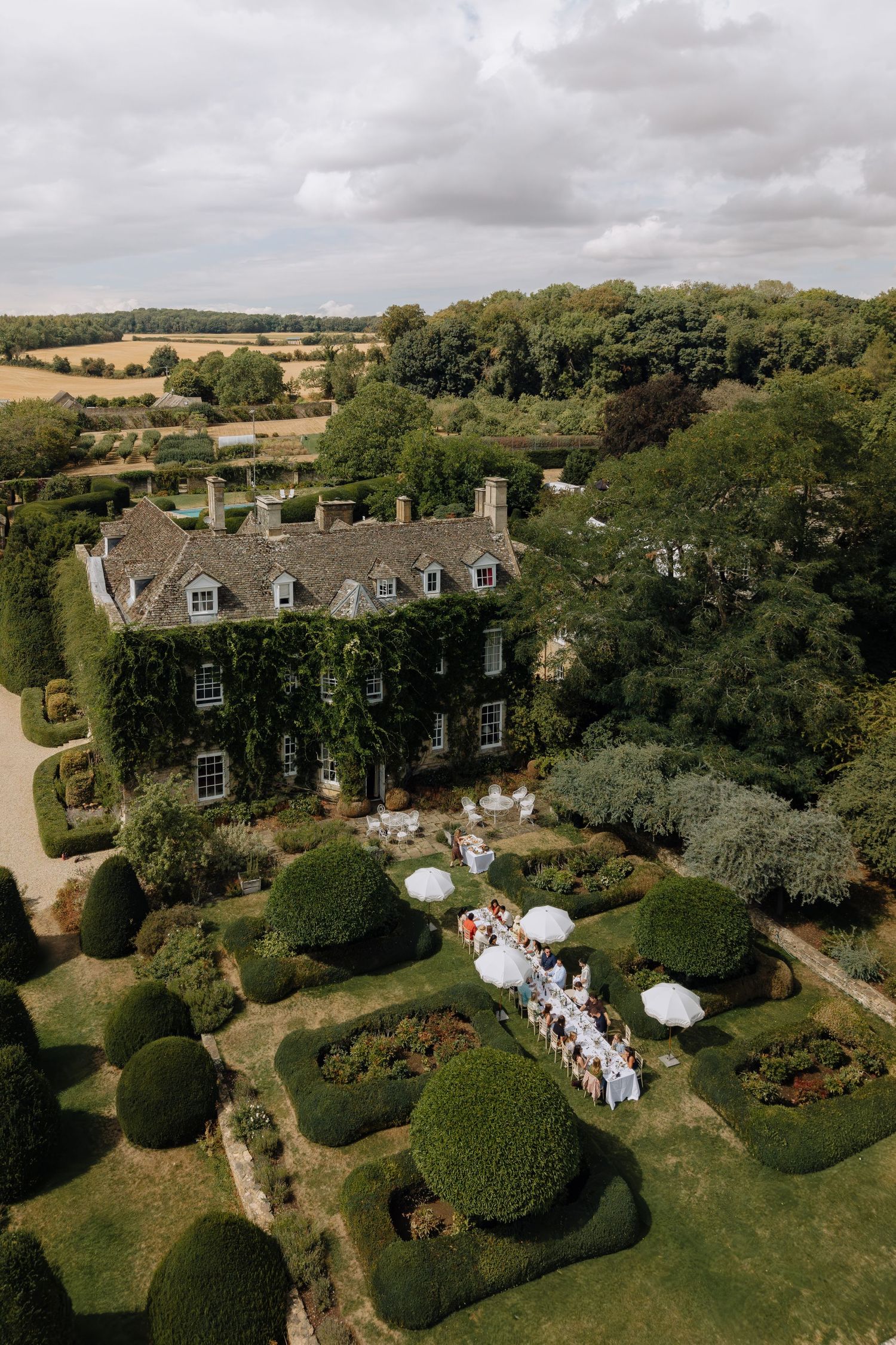 Aerial view of an ivy-covered English manor house with formal topiary gardens hosting an outdoor wedding reception.