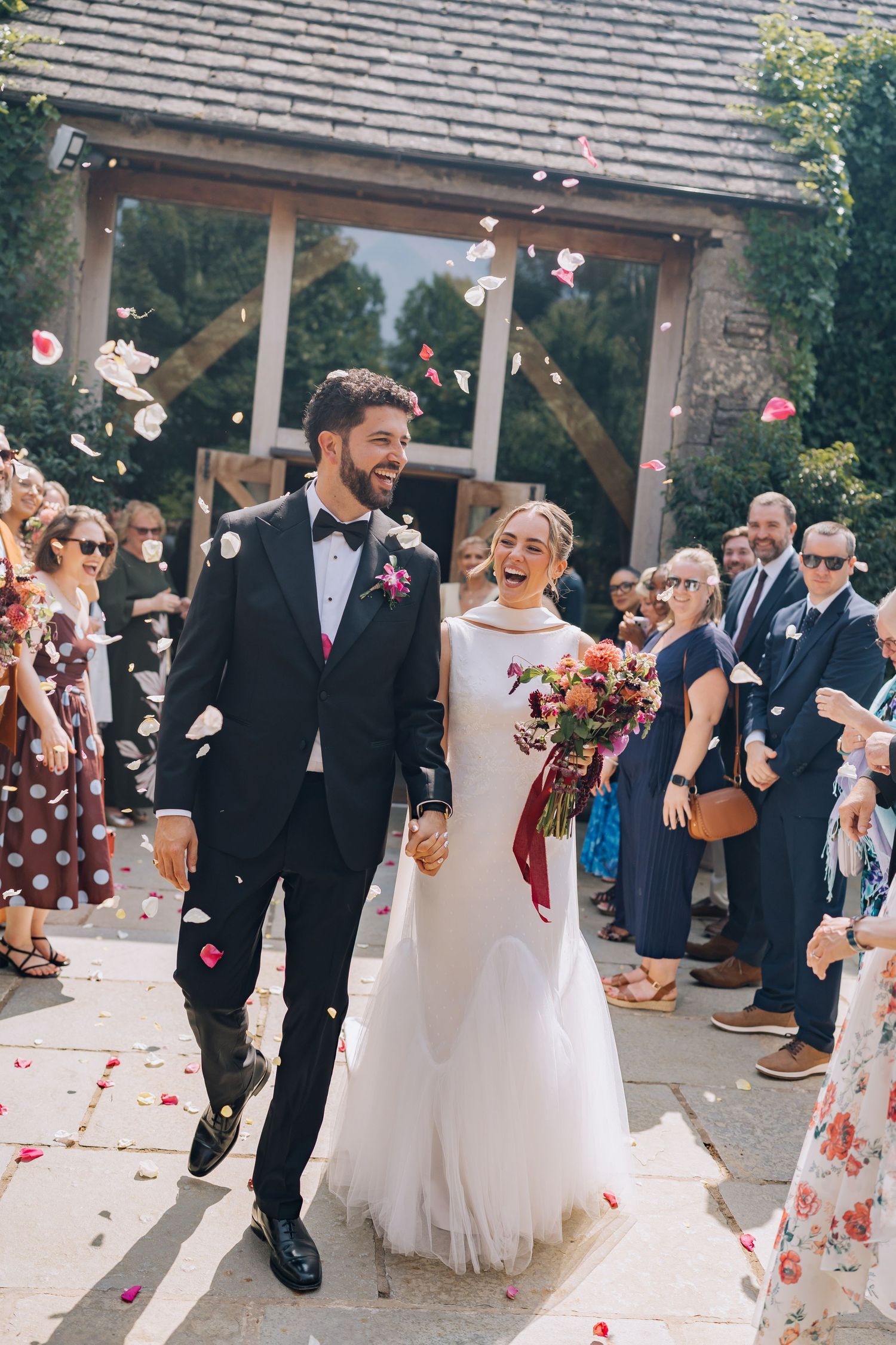 Joyful newlyweds walk through confetti shower outside rustic barn venue, bride holding colorful bouquet.