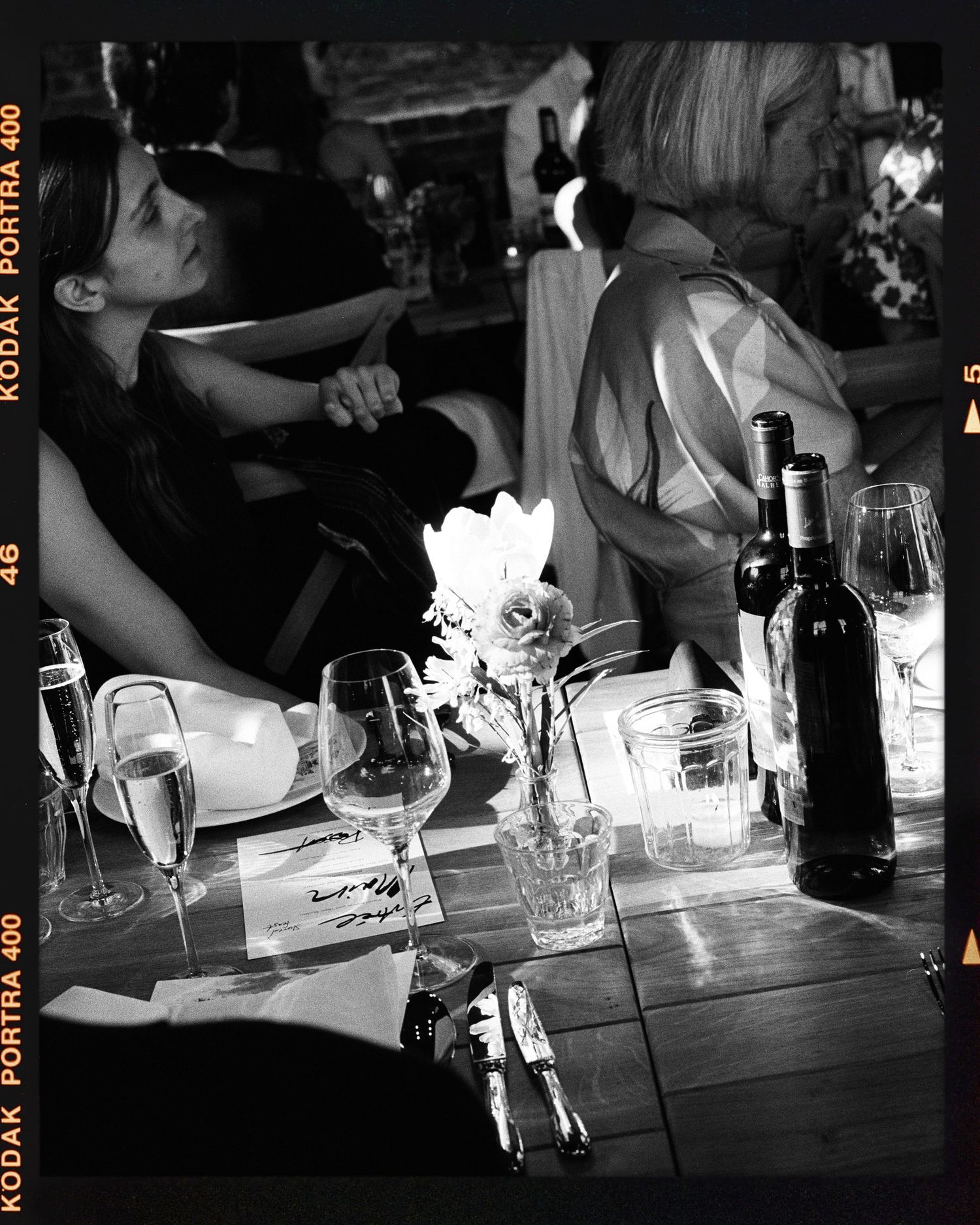 Black and white film photo of a woman at a dinner table with wine bottles and glasses at an elegant event.