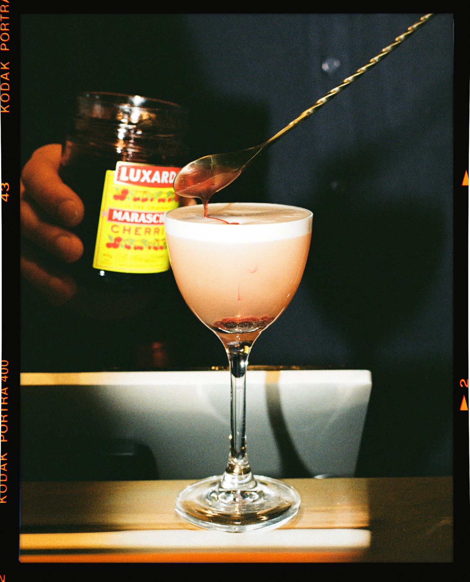 Bartender pouring Luxardo Maraschino cherry liqueur into a pink cocktail in a coupe glass with a bar spoon.