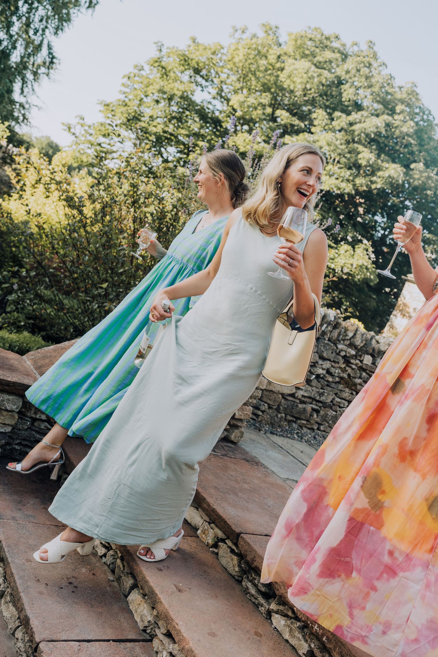 Three elegantly dressed wedding guests laughing and holding champagne glasses on stone steps outdoors.