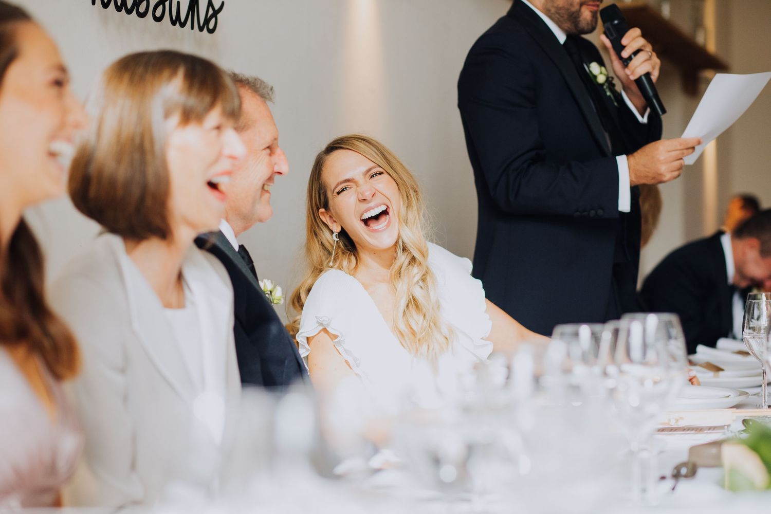 Bride laughing joyfully at wedding reception table during speeches in bright modern venue setting.
