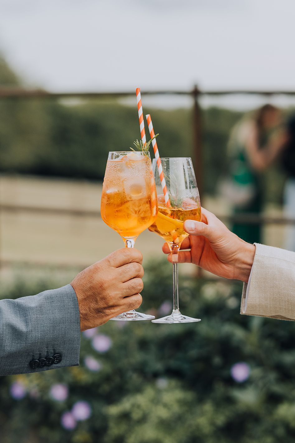 Two hands clinking glasses of orange Aperol Spritz cocktails at an outdoor wedding reception.