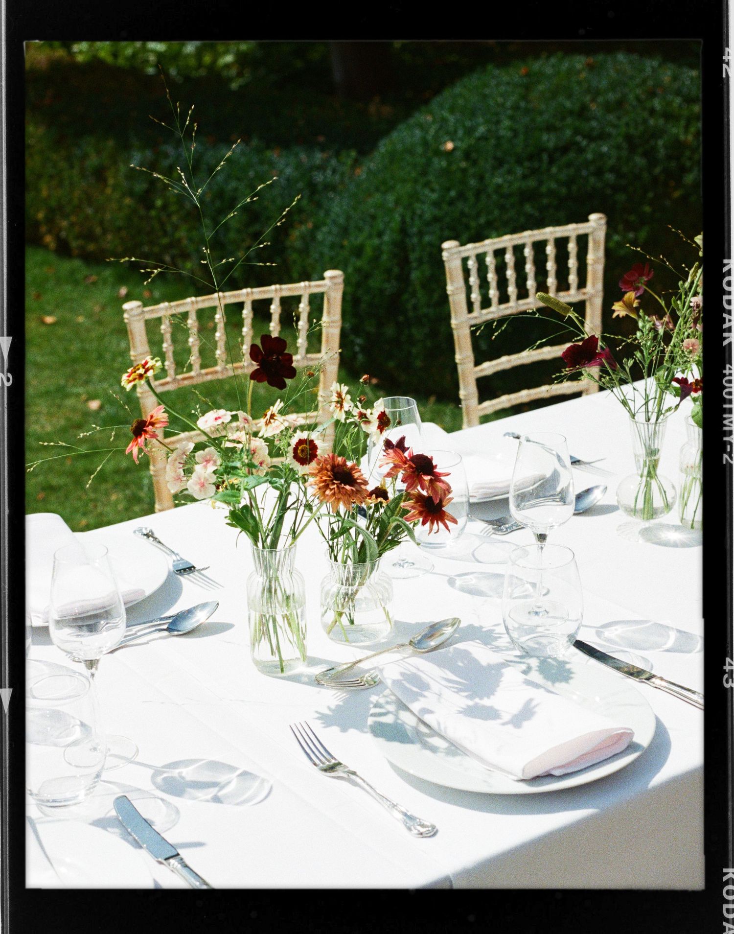Elegant outdoor garden dining table set with white linen, wildflower centerpieces, crystal glasses, and chiavari chairs.