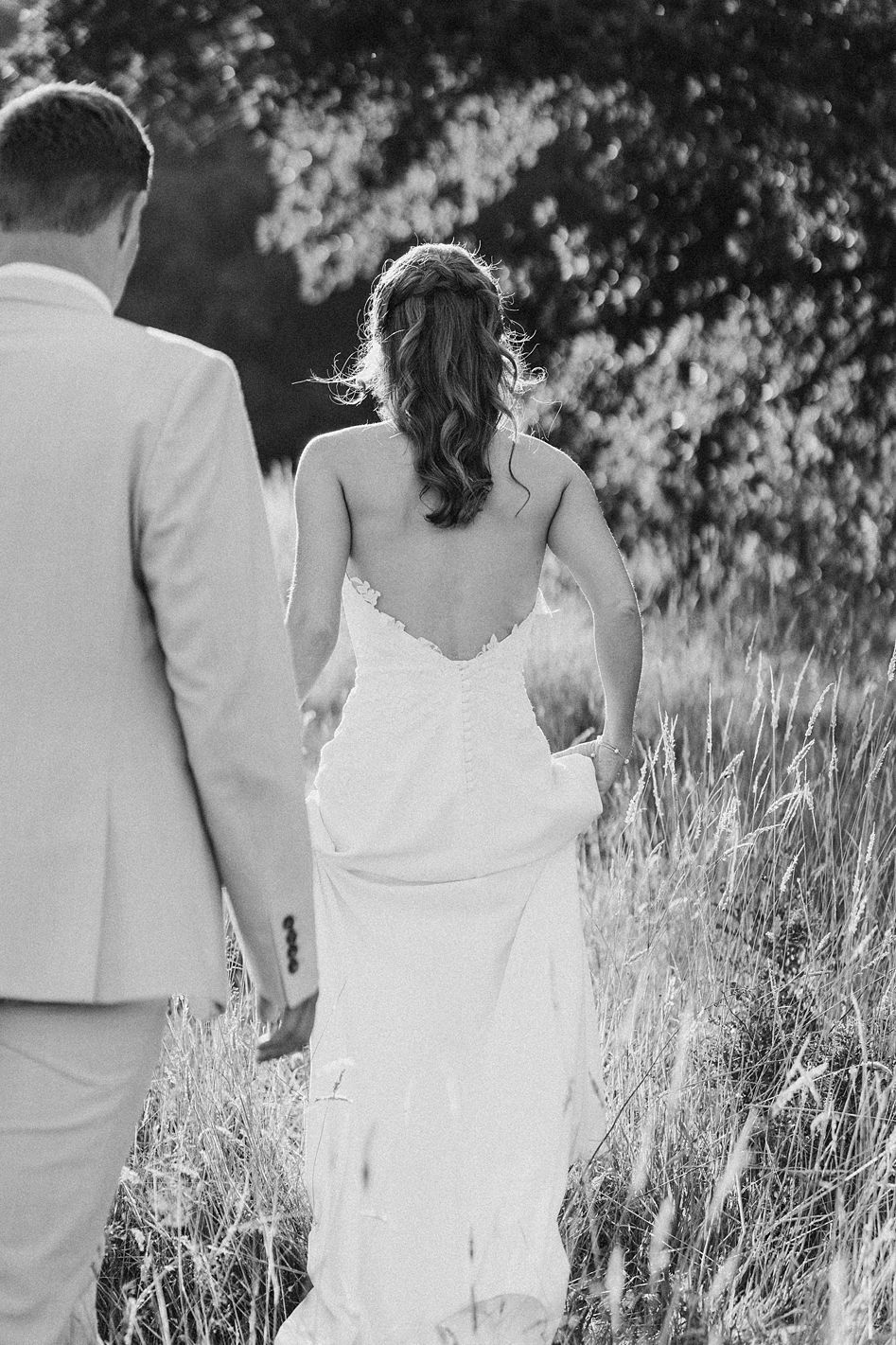 Black and white photo of bride in low-back lace gown and groom walking through tall grass meadow.