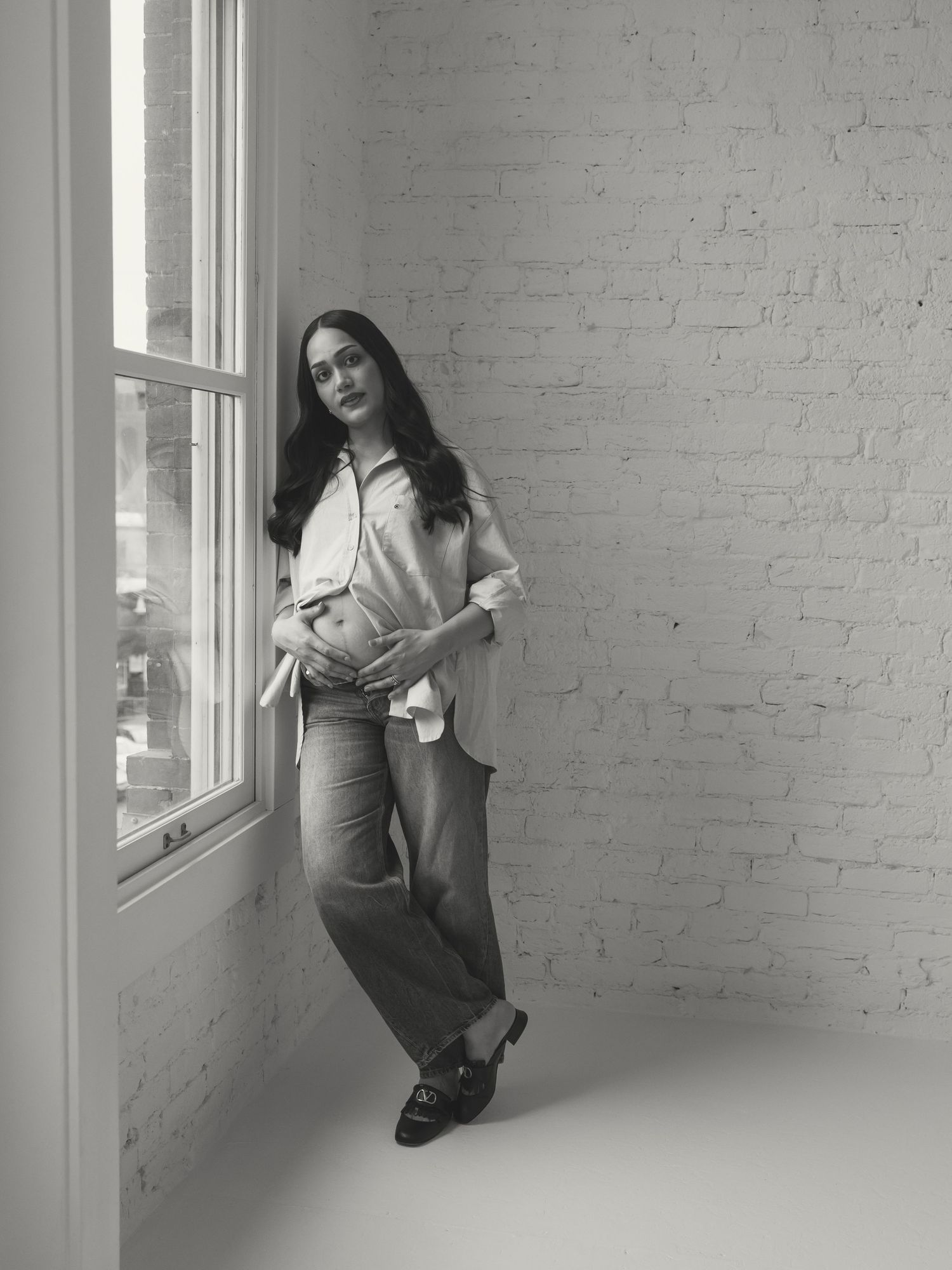 An expectant mother in casual attire leans against a white brick wall near a window.
