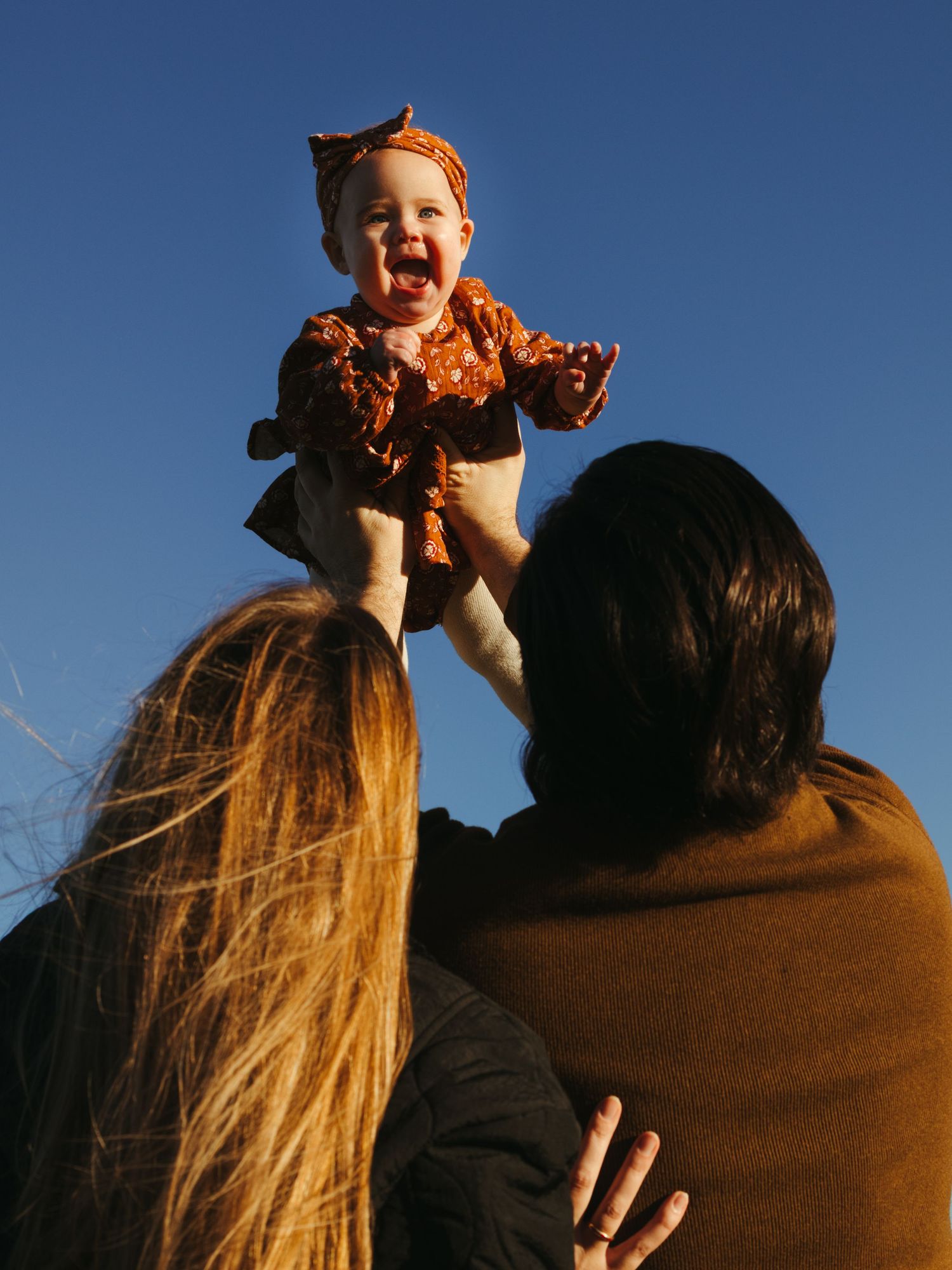 A joyful baby in orange clothing being lifted high against a bright blue sky.