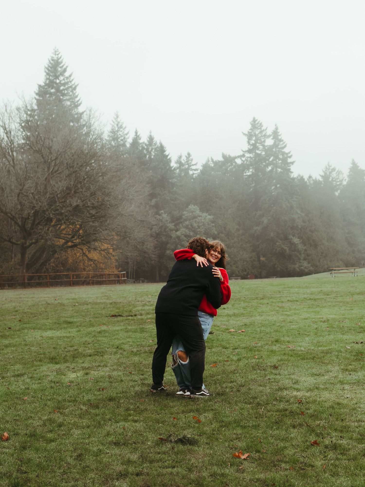 A couple shares an intimate moment in a foggy field with pine trees in the background.
