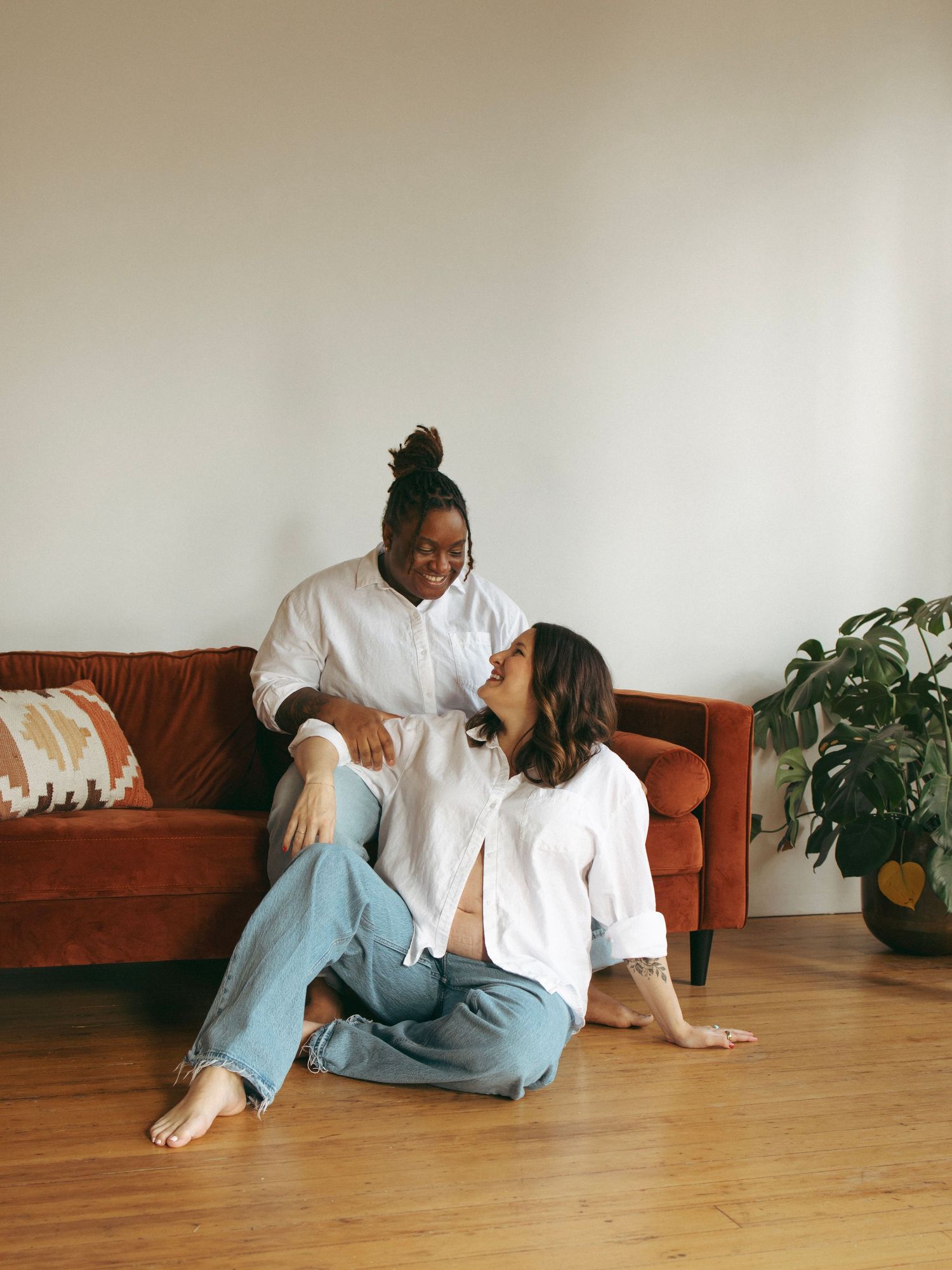 Two people relaxing on hardwood floor near brown couch and houseplants in casual setting.
