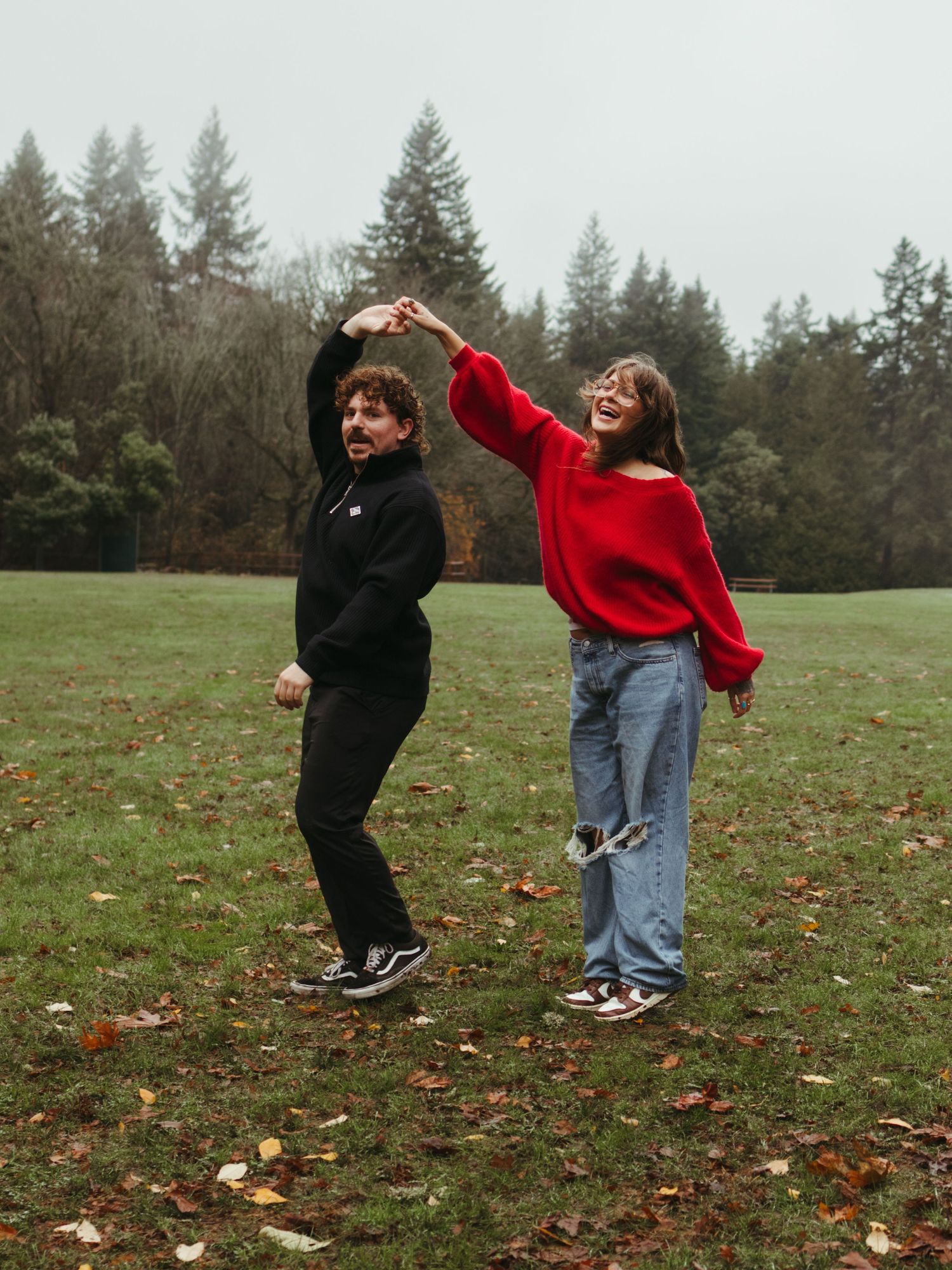 A couple dances and spins playfully in a grassy field on an overcast day.