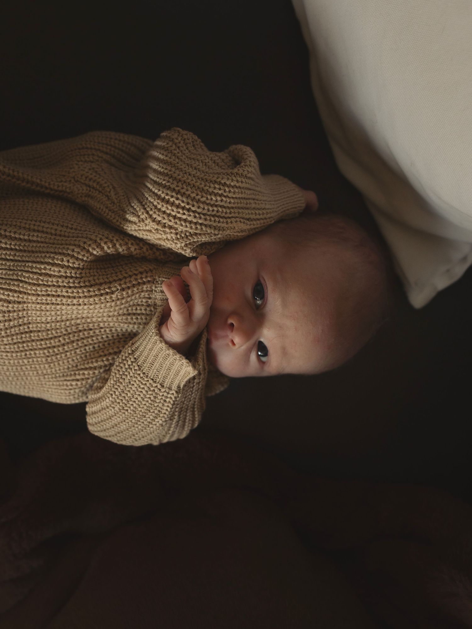 A baby wearing a knit sweater looks up at the camera in moody lighting.