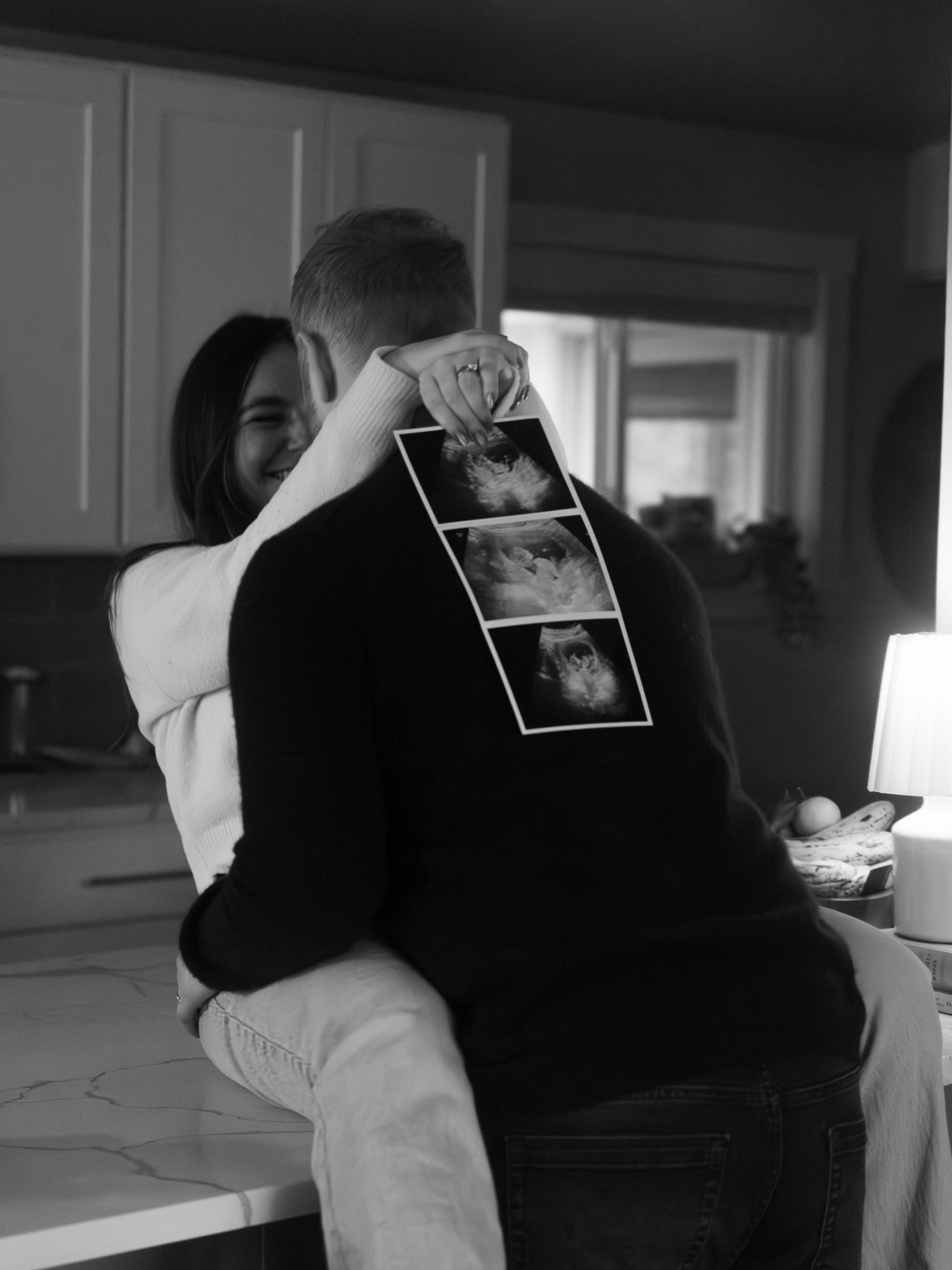 A couple sharing an embrace on a kitchen counter while holding sonogram images.