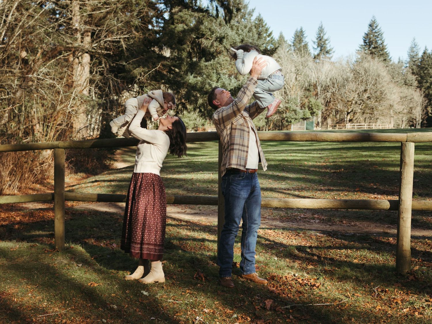 A couple plays with their child in a scenic outdoor setting surrounded by evergreen trees.