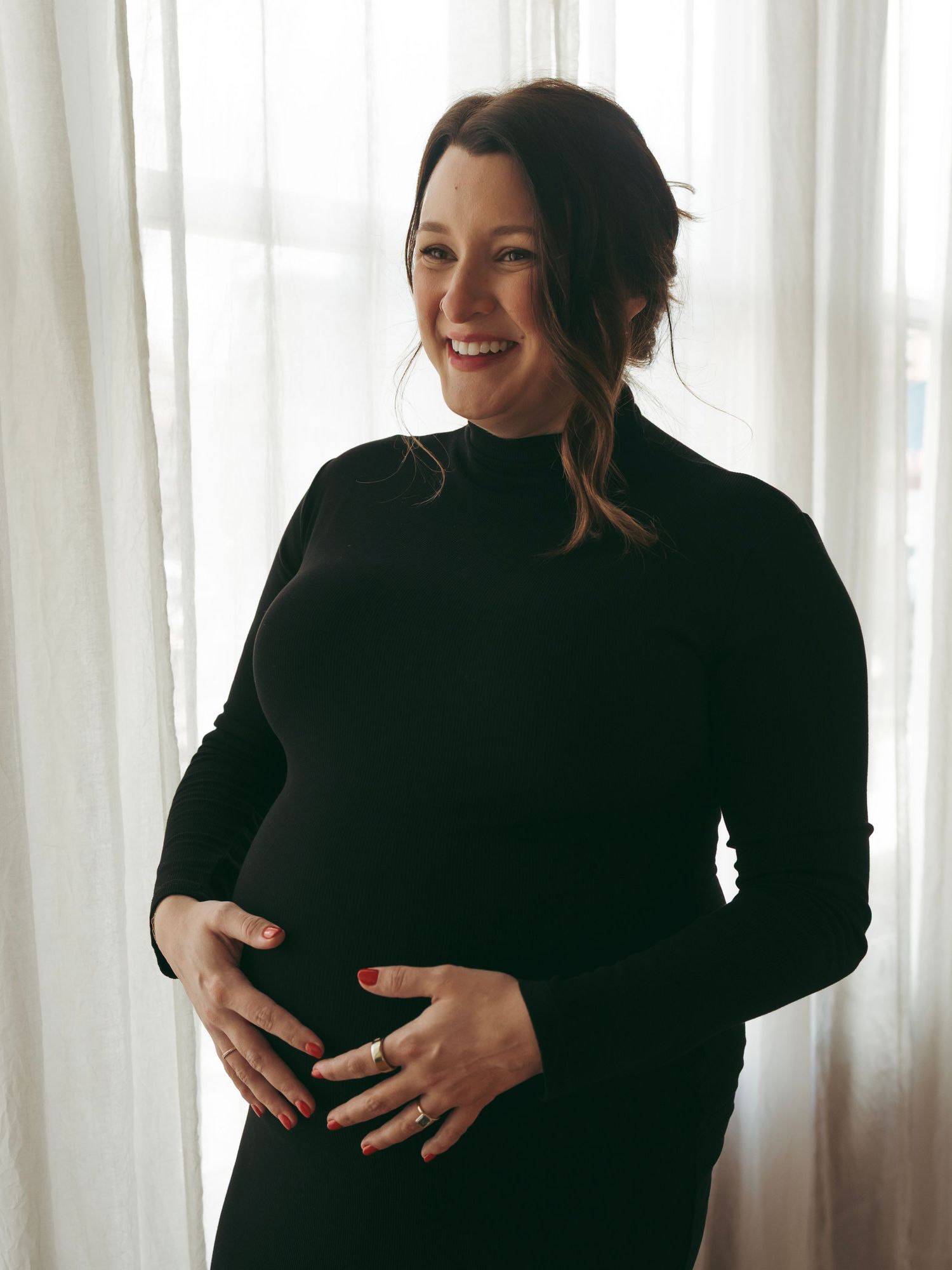 Maternity pose in black sweater poses against white curtains in natural window light.