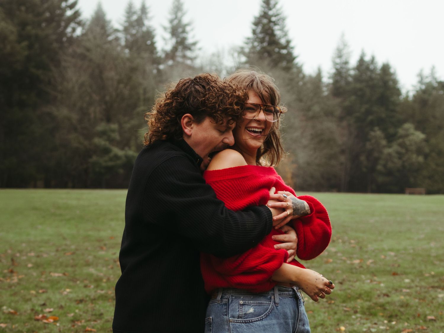 A couple embraces joyfully in a misty field surrounded by evergreen trees.