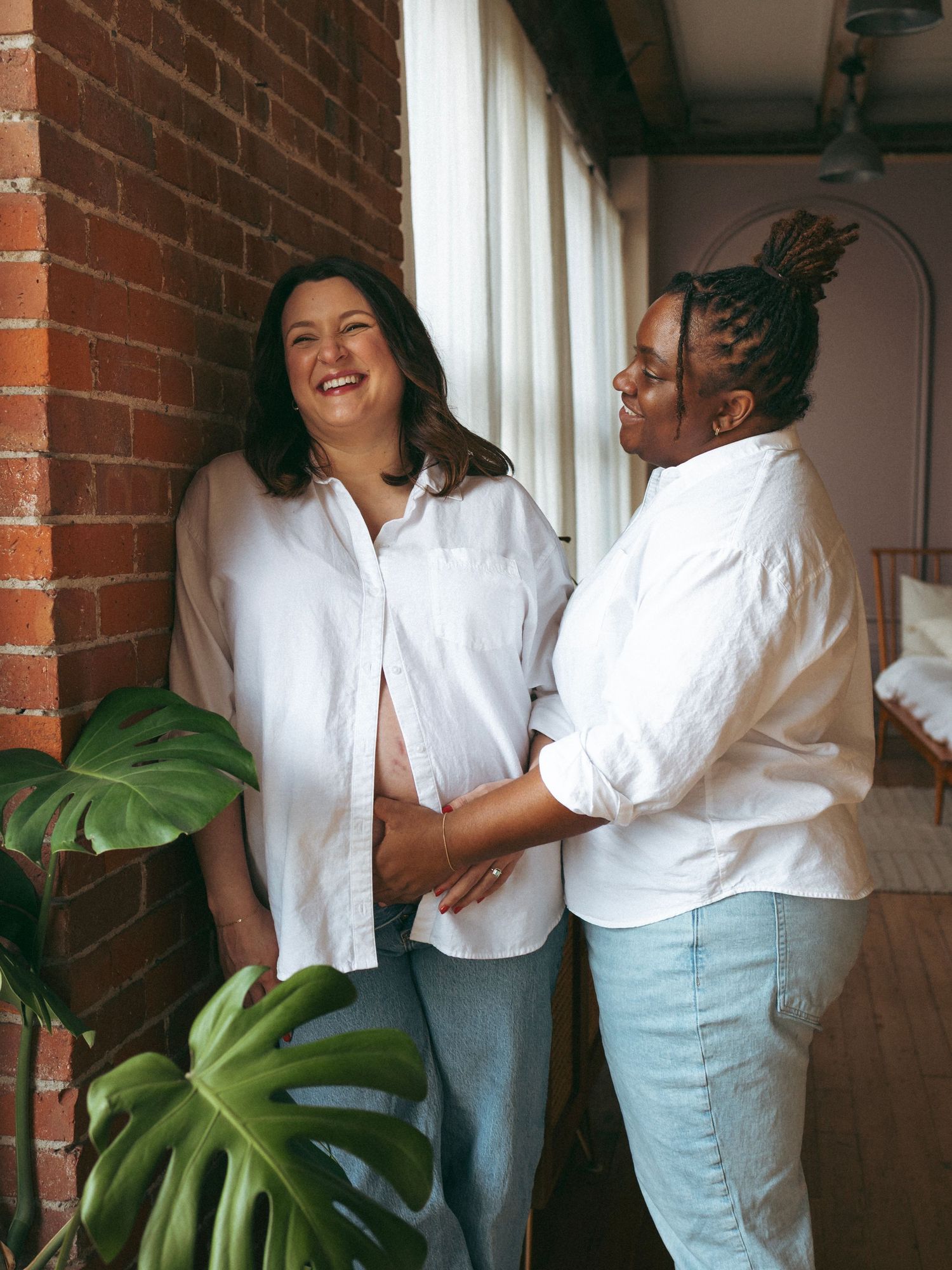 Two women in matching white shirts and jeans share joyful moments against a brick wall backdrop.