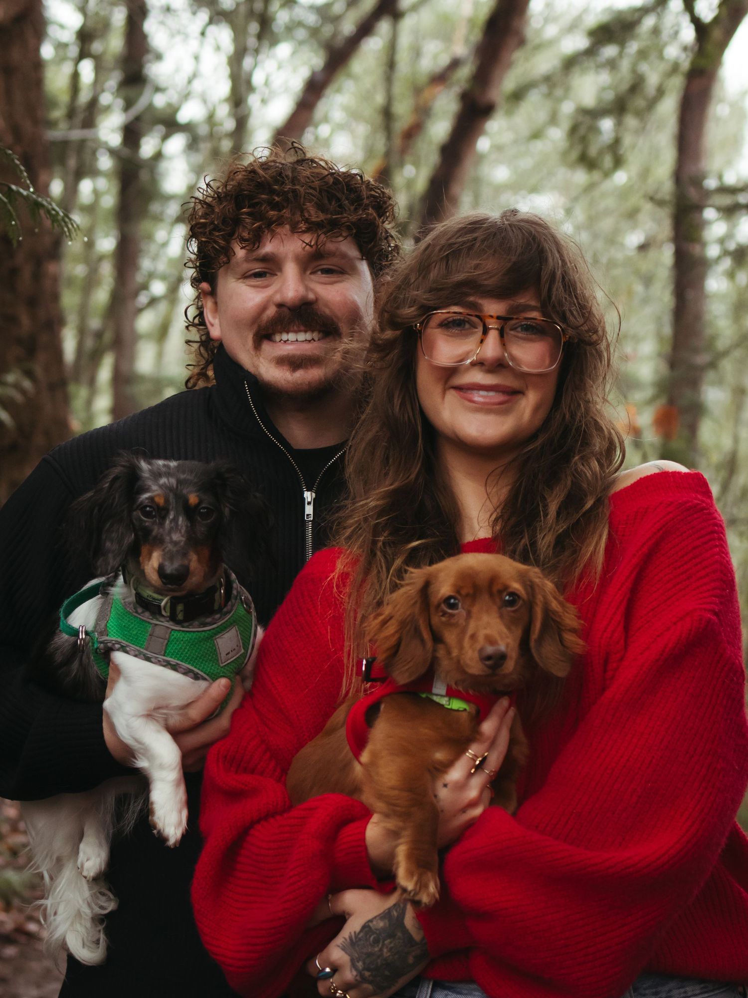 A couple poses with their small dogs in a wooded setting wearing red and black outfits.