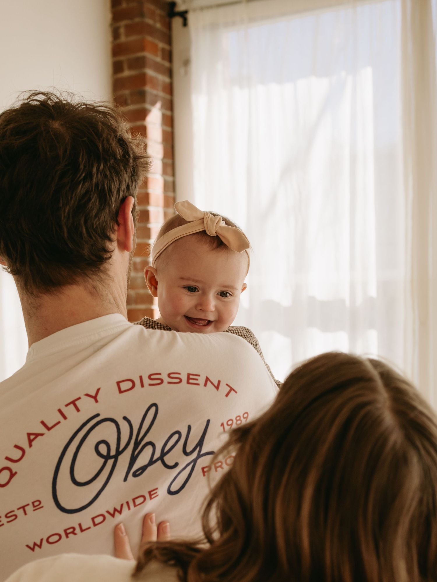 Bright indoor family photoshoot with sunlit windows and exposed brick wall in background.