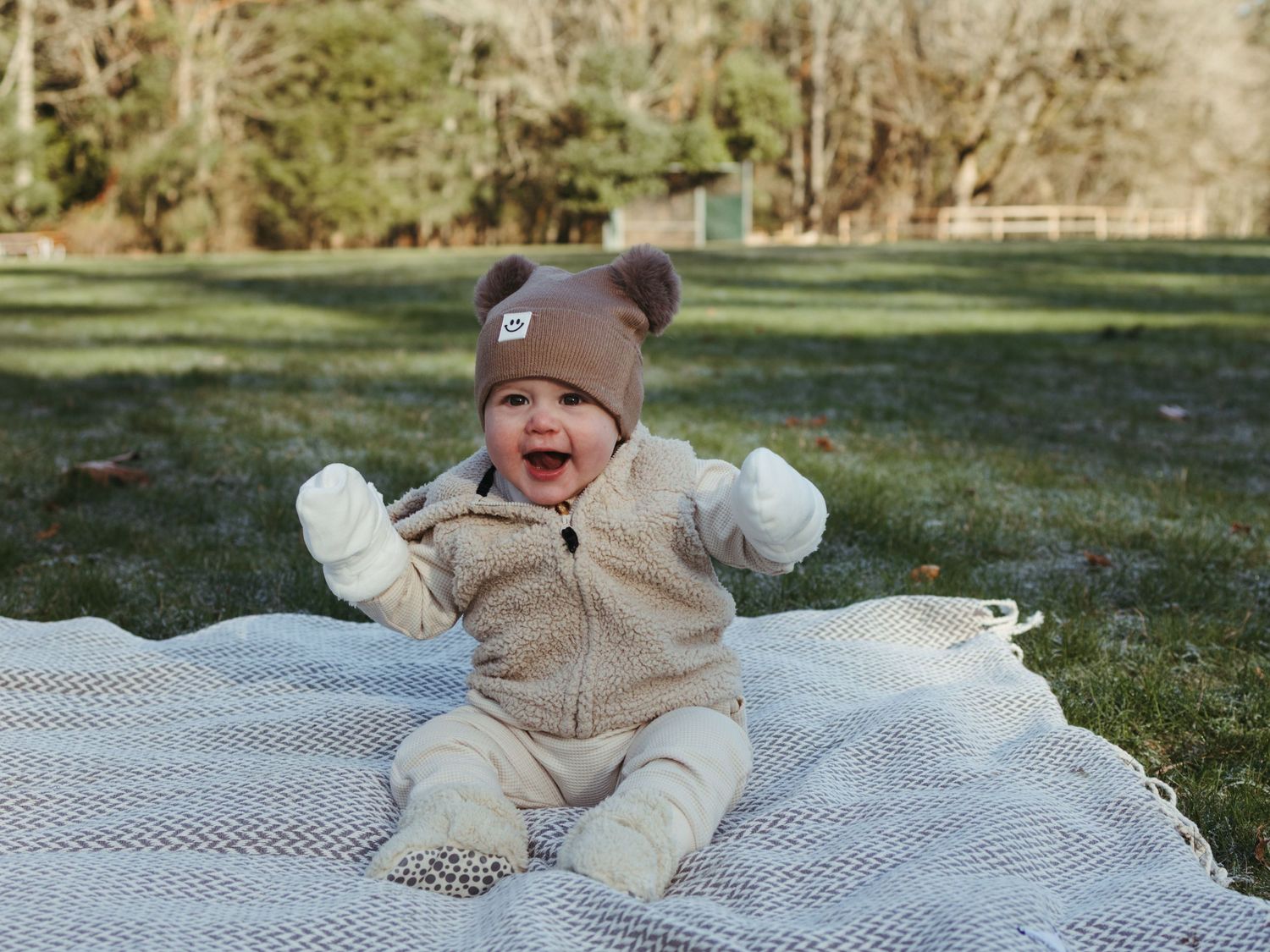 A baby in a bear hat sits on a blanket outdoors expressing joy.