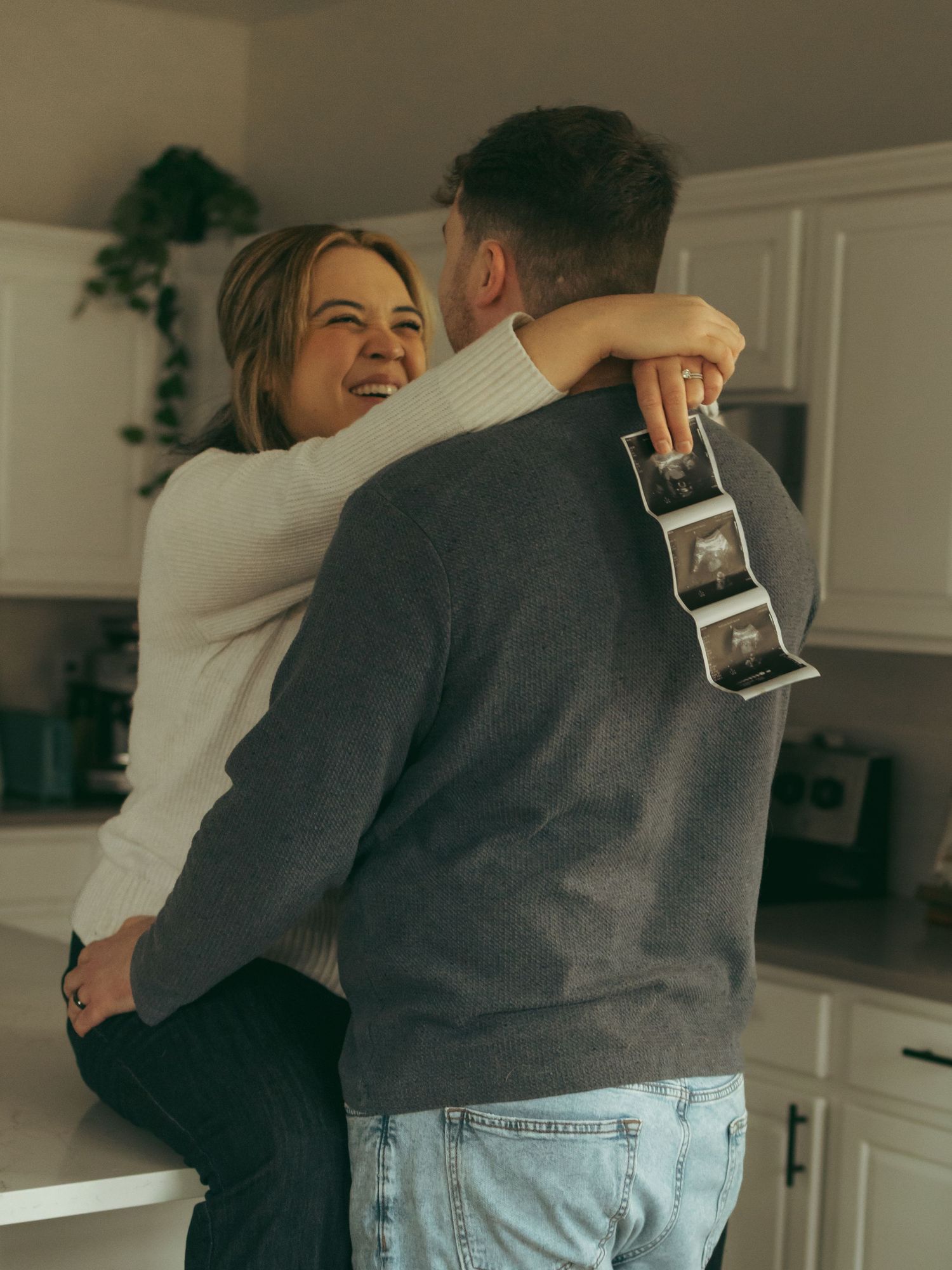 A joyful couple embracing in their kitchen while holding ultrasound pictures.