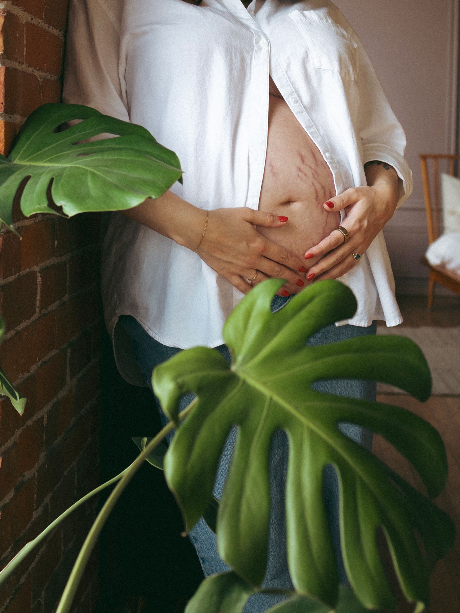 Hands gently touching pregnant belly near a large green monstera leaf against brick wall.