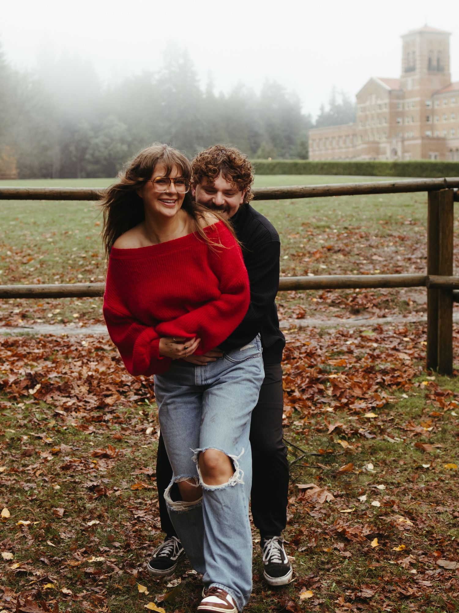 A couple poses playfully by a wooden fence surrounded by fallen autumn leaves.