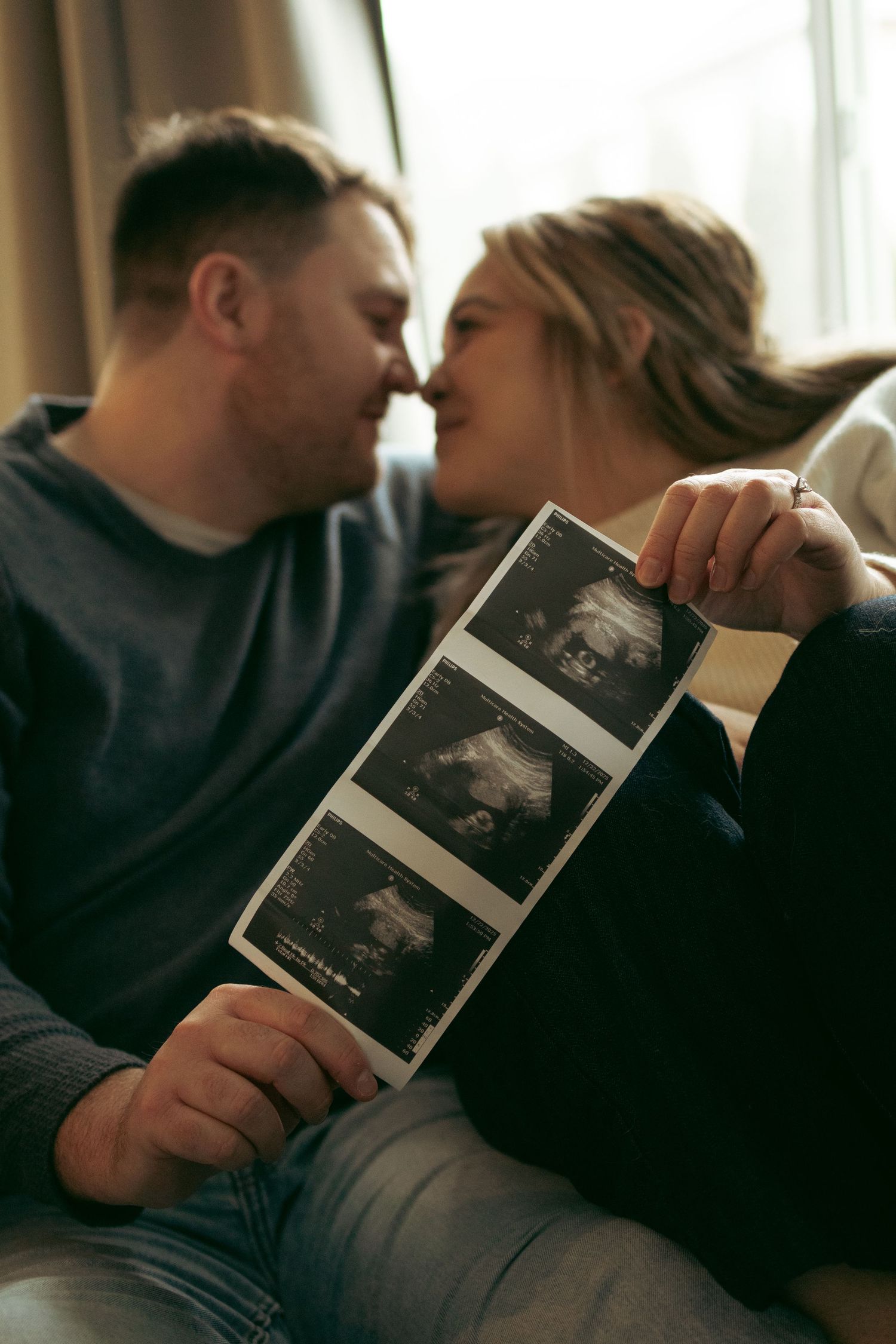 A couple sharing an intimate moment while looking at ultrasound photos together on a couch.