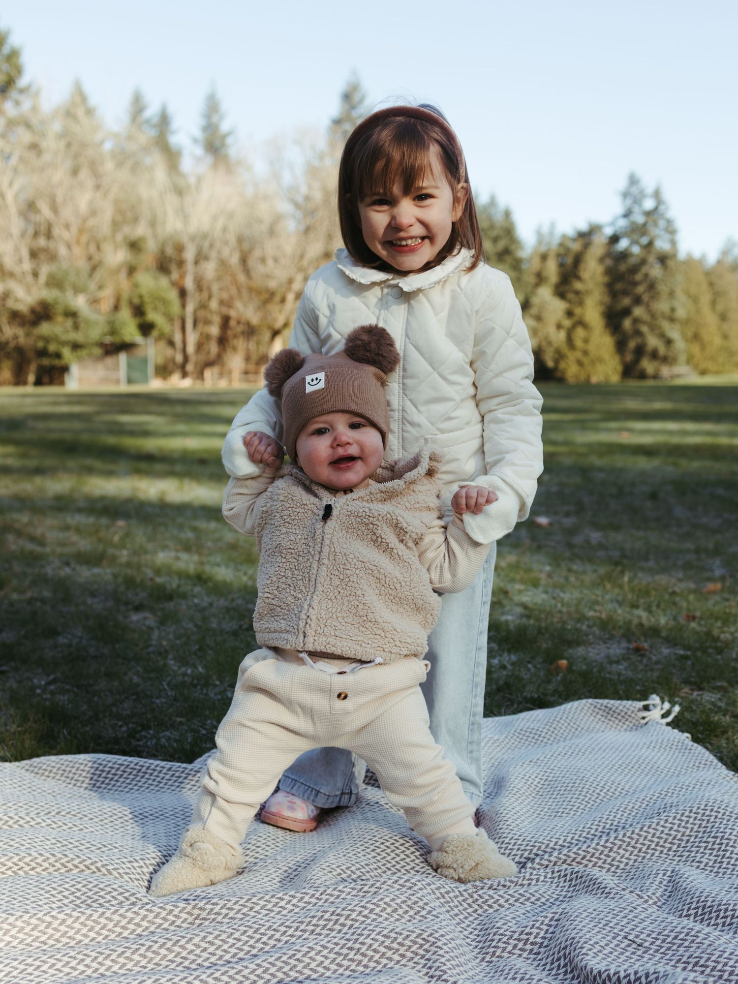 Two children in warm winter clothing play together on a picnic blanket outdoors.