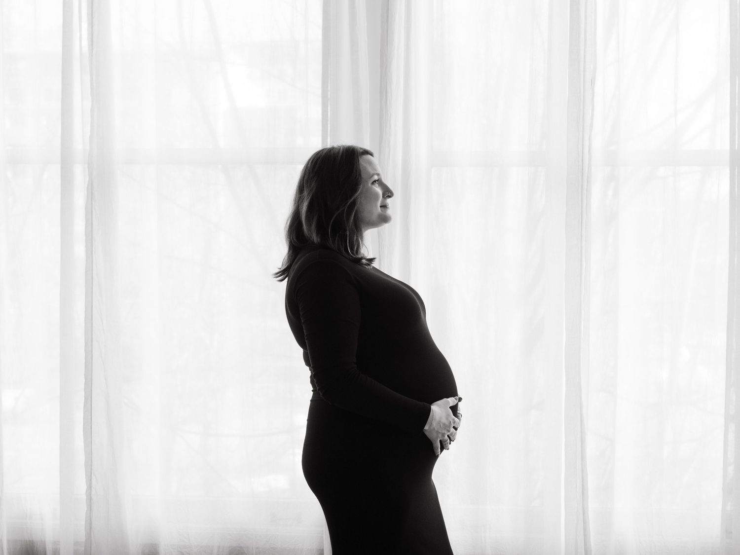 Pregnant woman silhouettes stand against bright window light in an artistic black and white composition.