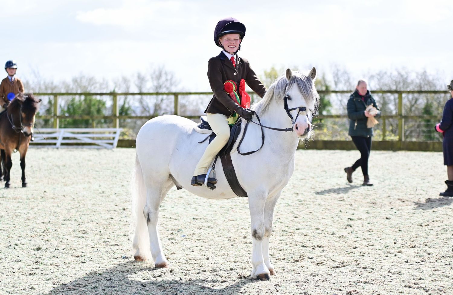 Equestrians in riding gear compete in a show jumping event on a sandy outdoor arena.