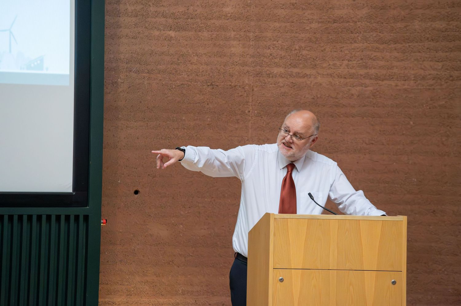 Speaker presenting at podium in modern conference room with wood paneled walls.