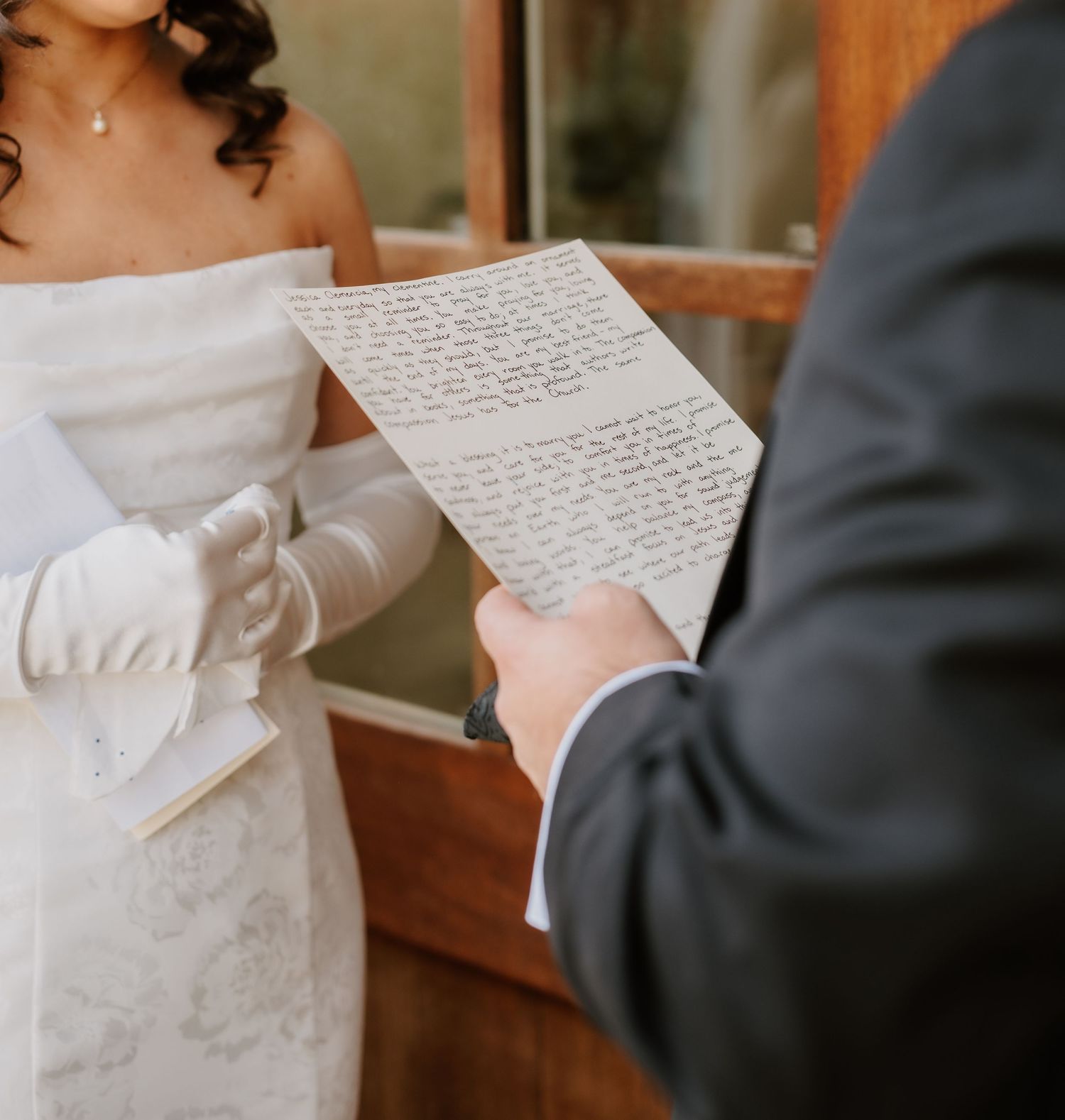 Groom reading handwritten wedding vows letter while bride in white gloves listens during first look.