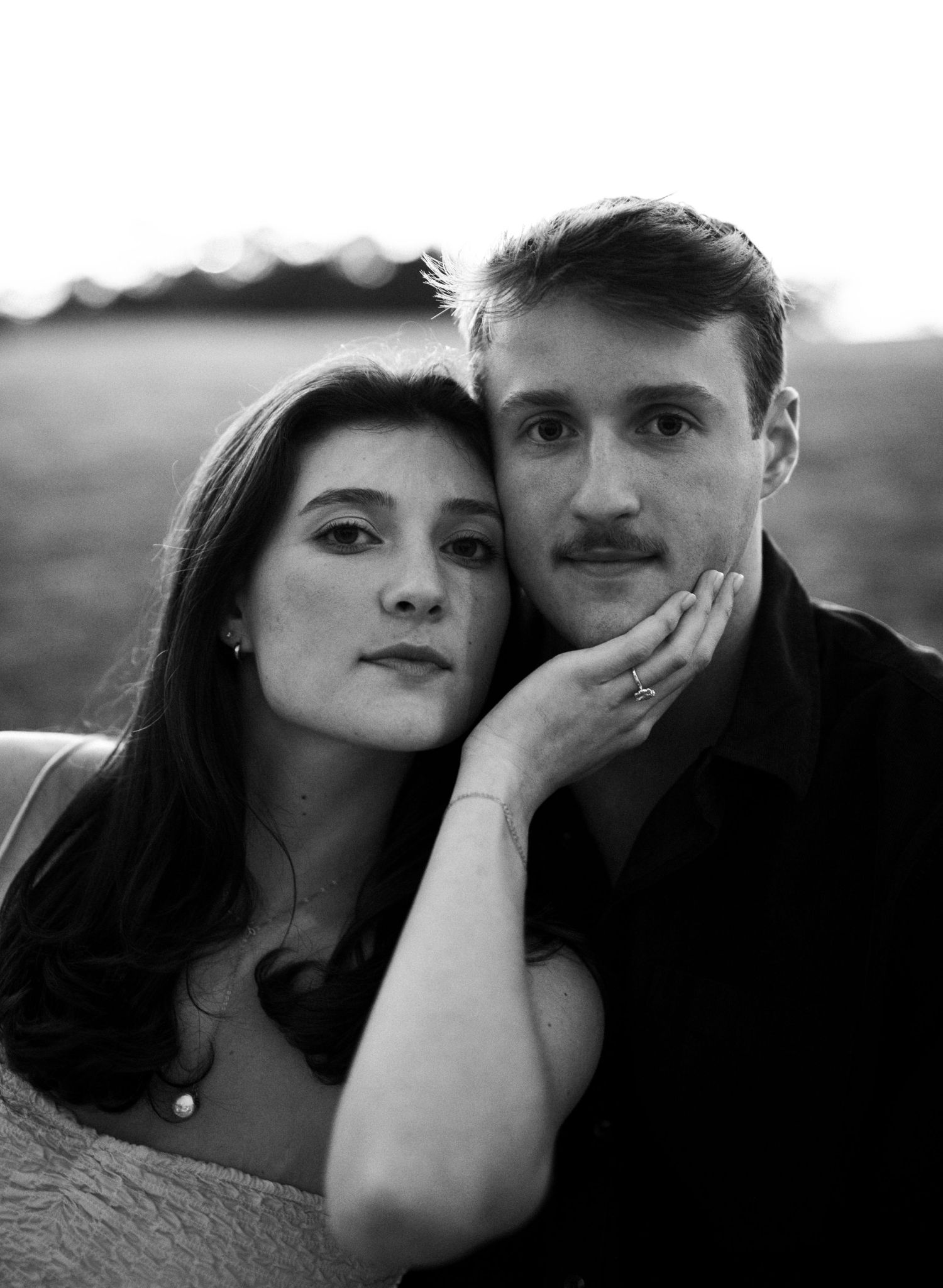 Black and white portrait of engaged couple, woman resting head on mans shoulder in soft evening light.
