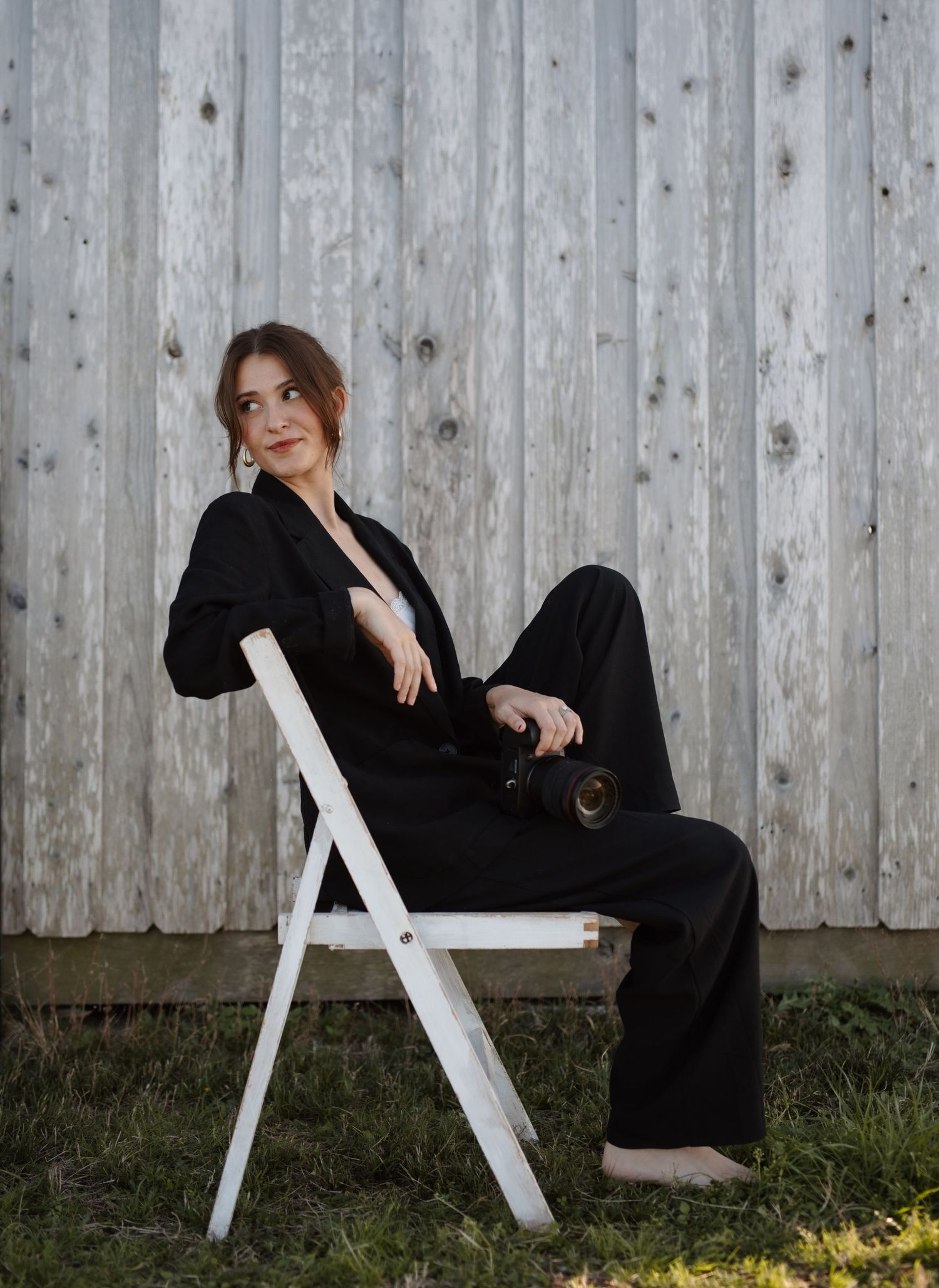 Female photographer in black suit sitting on white folding chair holding camera by wooden fence.