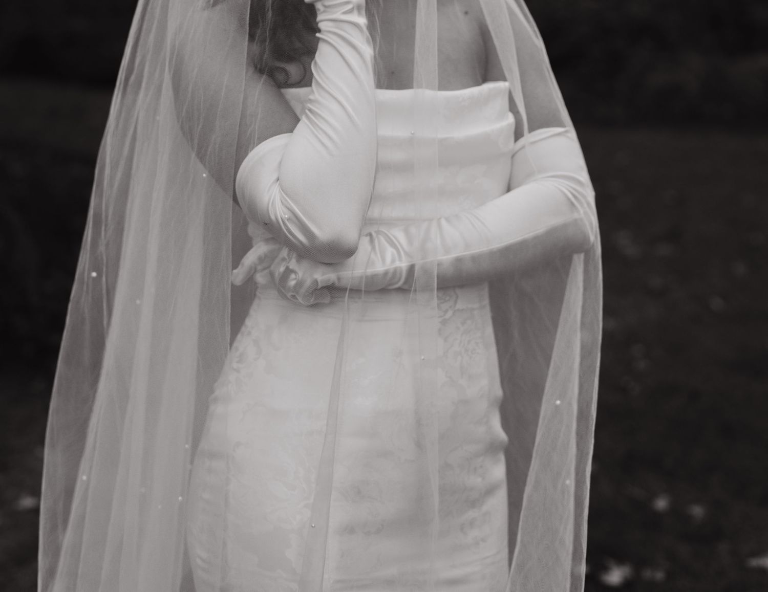 Black and white portrait of bride in fitted gown, long gloves, and flowing cathedral veil.