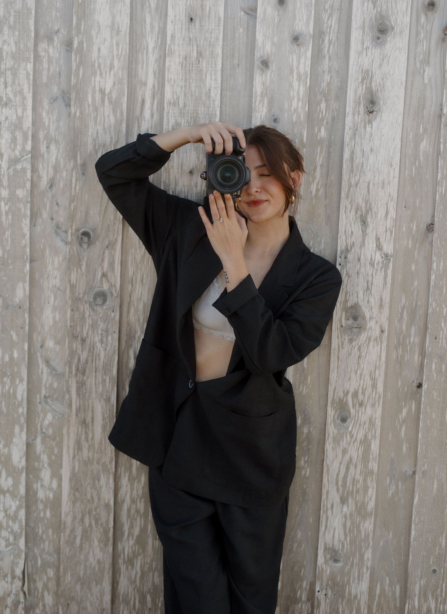 Woman in black suit and white crop top taking photo with DSLR camera against wooden plank wall.
