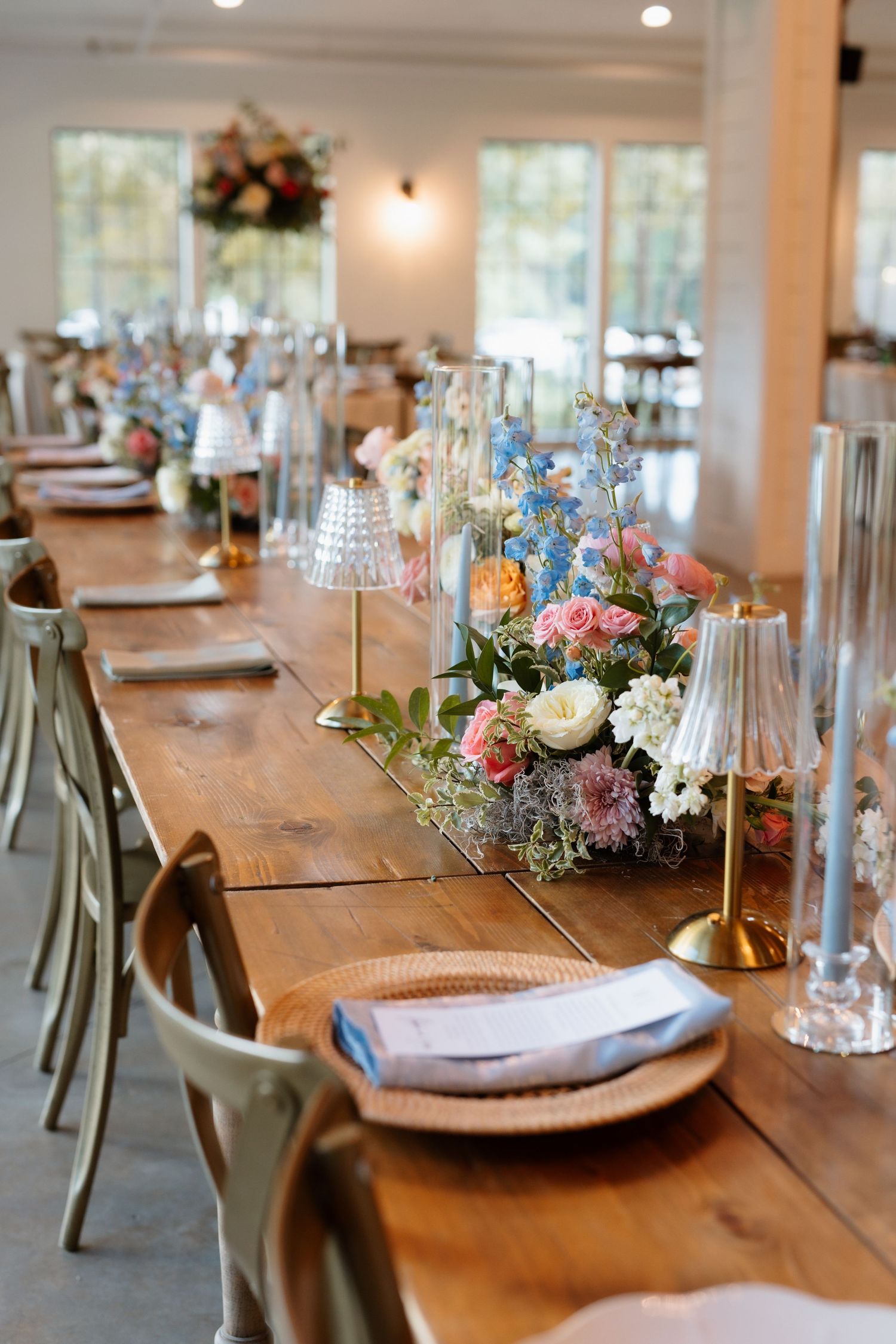 Elegant wedding reception table with floral centerpieces, gold candle holders, wicker chargers, and sage green chairs.