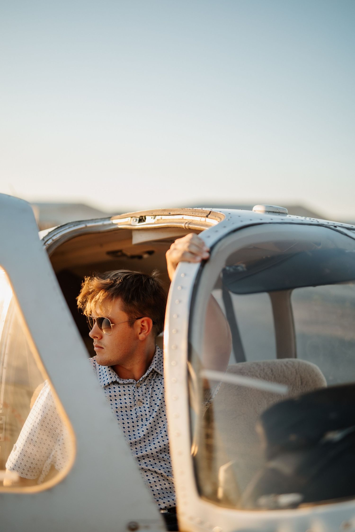 Young male pilot in sunglasses exiting a small single-engine aircraft cockpit at golden hour.