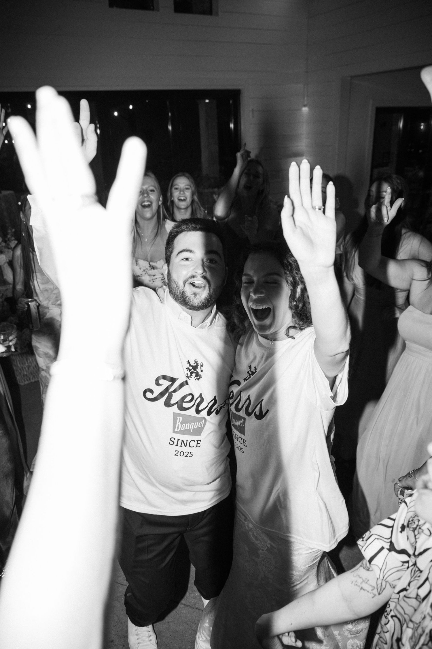An excited couple in matching Kerrs wedding t-shirts celebrate with hands raised on the dance floor.